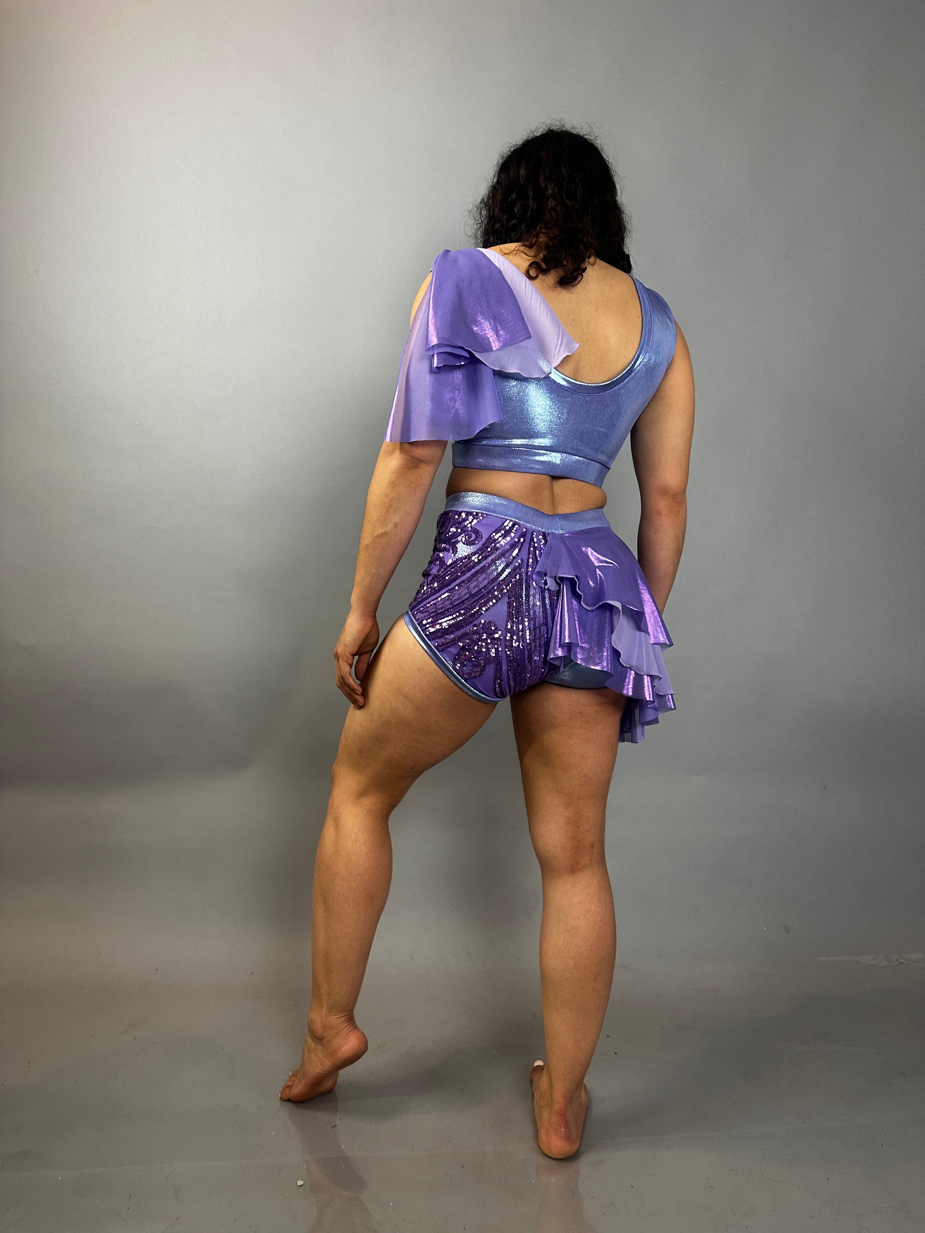 A person stands barefoot facing away, wearing a lavender purple Sequin Dance Costume featuring high waist shorts with half skirt, a crop top, and 4-way stretch mesh.