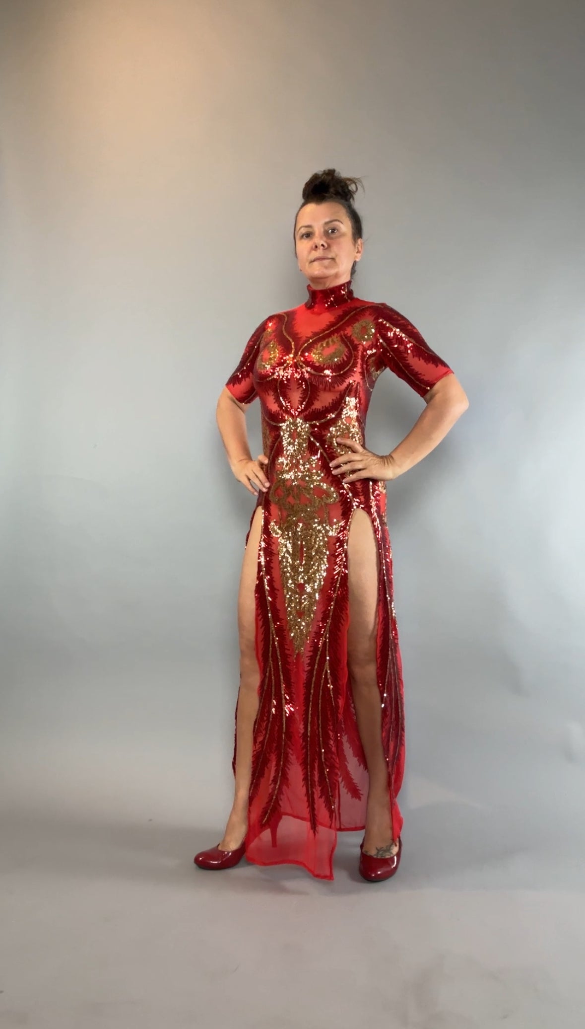 A woman stands confidently against a gray backdrop in the Red And Gold Sequin Sheer Sleeveless Dress, styled with hand accessories.