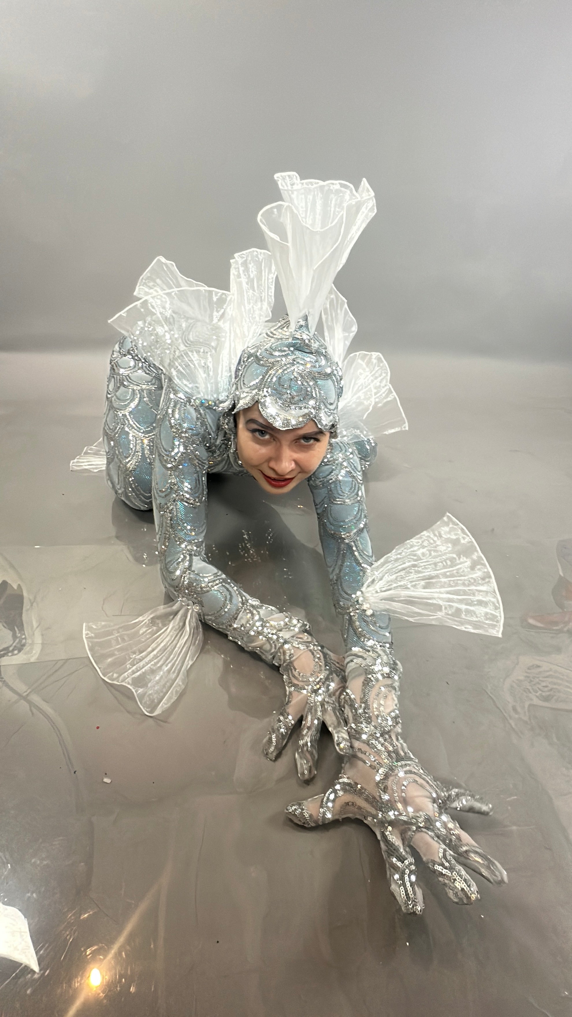 A person in the Sea Creature costume crouches on the floor, displaying translucent fins and silver scales in this striking wearable sculpture.