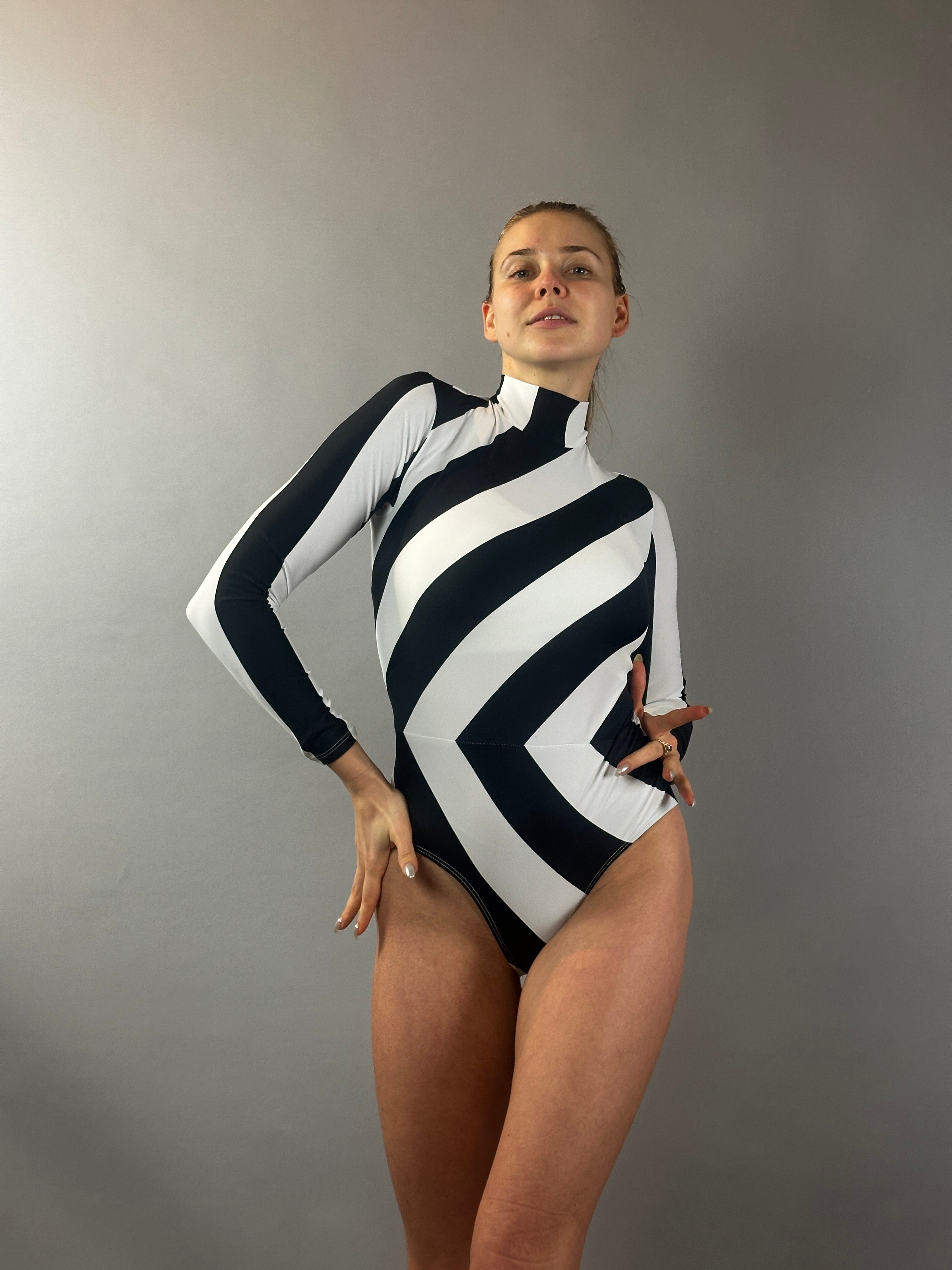 Model wearing the Black and White Striped Leotard— a mock neck, long sleeve geometric print bodysuit—poses against a plain gray background. Perfect as an aerialist outfit, gymnastics wear, or dance costume.
