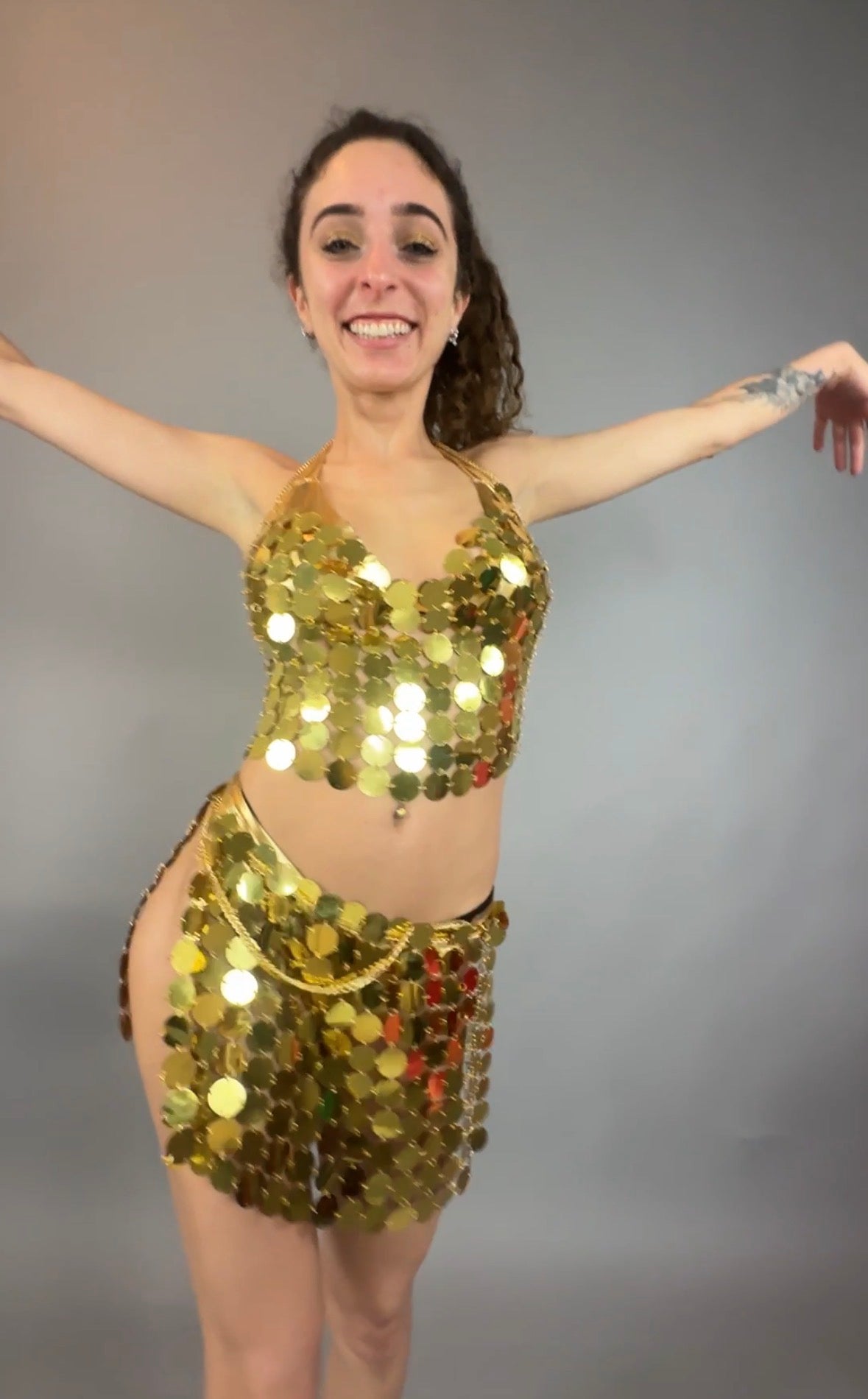 A woman smiles with arms raised, modeling the Gold Chain Two-Piece Set for Performers costume, featuring a supportive crop top with gold sparkle against a plain gray background.