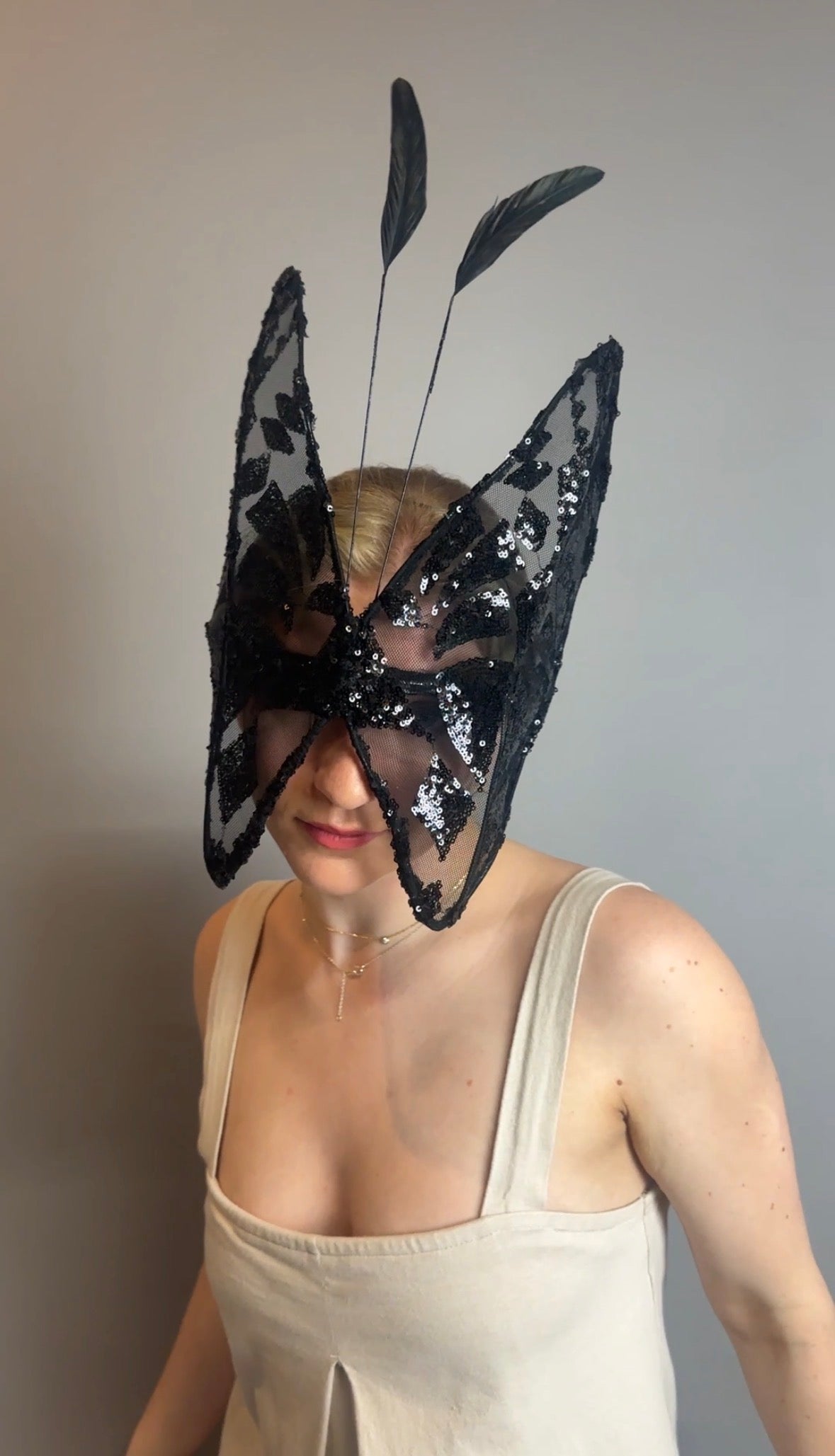 A woman wears the Butterfly Headpiece, a black lace hat with antennas and an open face, as a festive holiday costume accessory, standing against a plain gray background.
