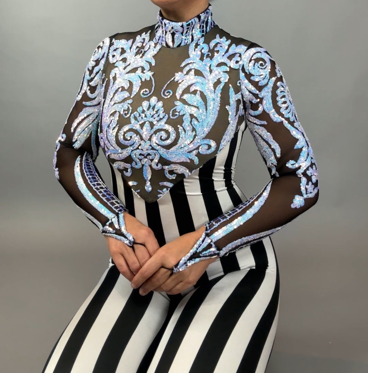 Model wears the Glamorous Sequins Stripes catsuit costume, featuring a sparkly sheer top with sequined leaf design and black and white vertical stripes, posed against a gray background.