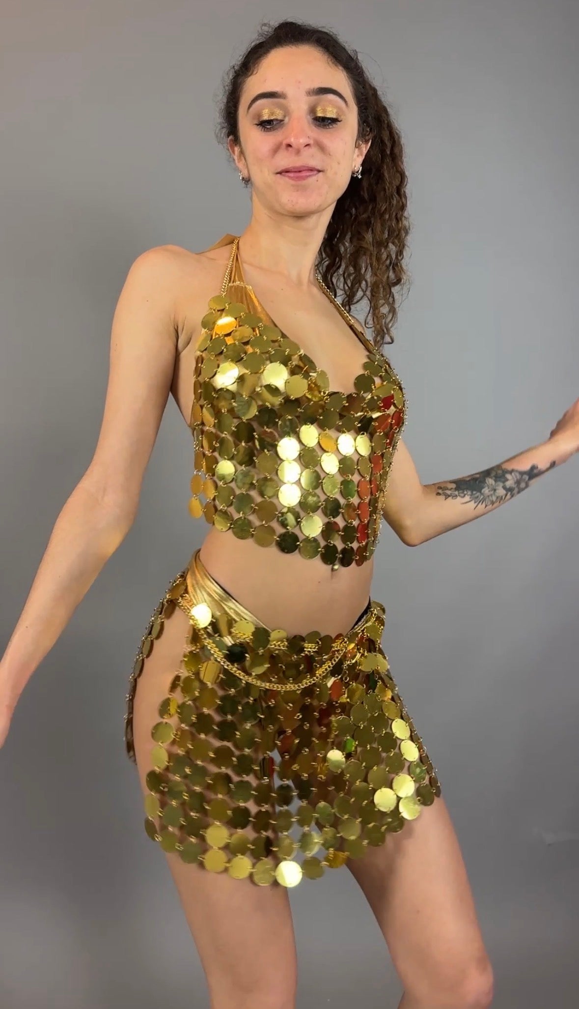 A woman models the Gold Chain Two-Piece Set for Performers—featuring a sequin halter top and skirt—posing confidently against a plain gray background.