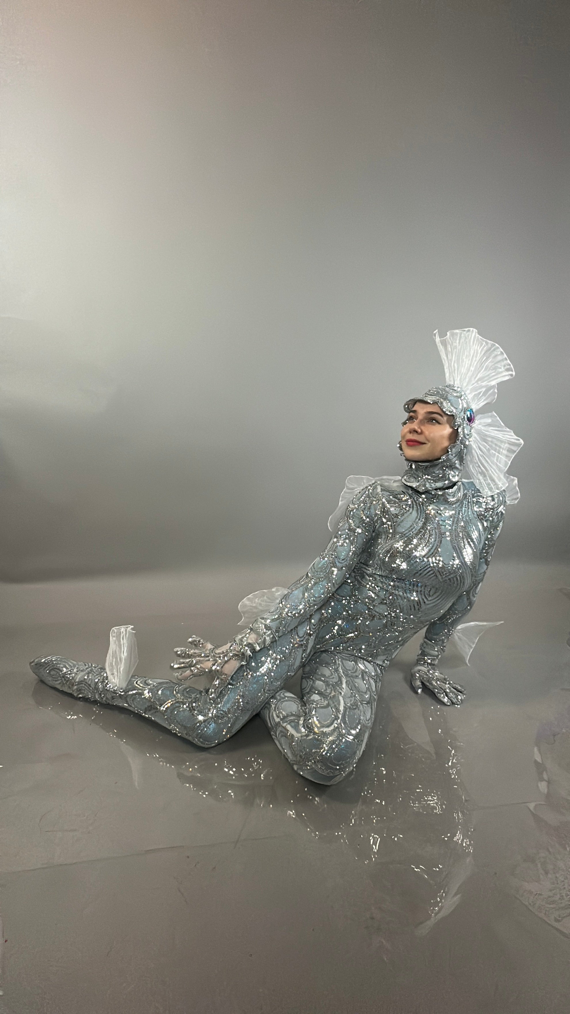 Model in the Sea Creature bodysuit with an elaborate headpiece poses seated on the floor against a gray backdrop, capturing the striking allure of couture-inspired design.