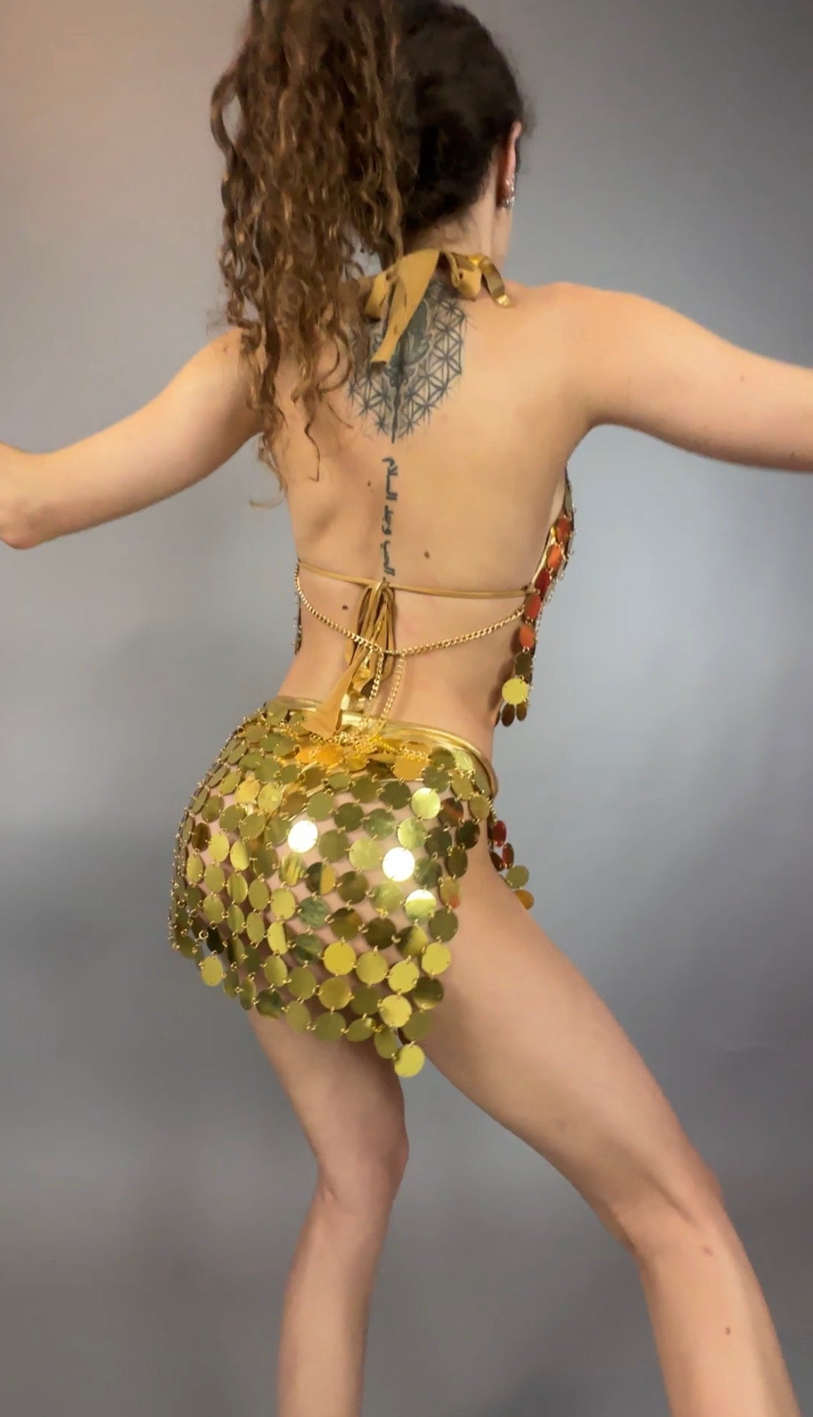 Dressed in the Gold Chain Two-Piece Set for Performers, a person poses with arms outstretched, showcasing a geometric tattoo on their upper back.