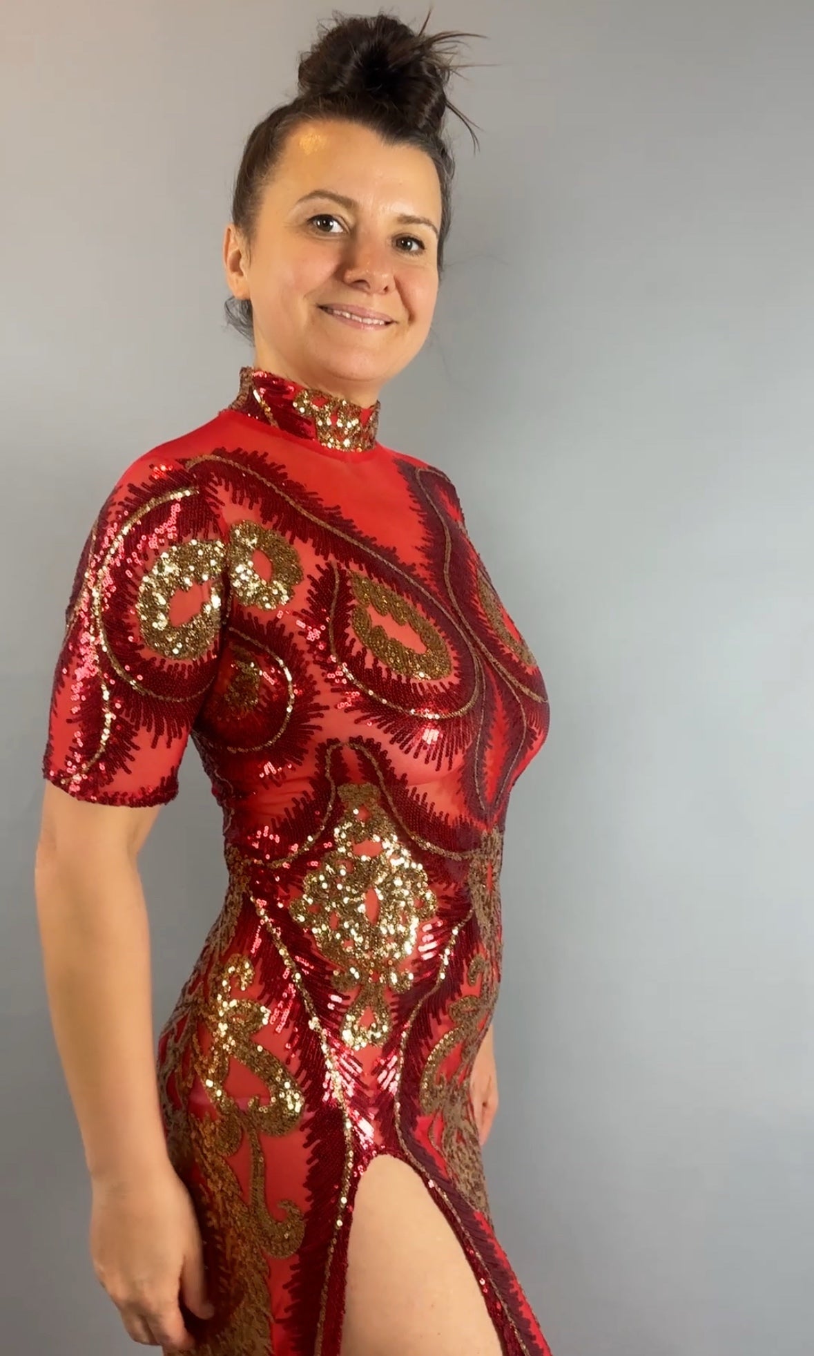 Woman wearing a Red and Gold Sequin Sheer Sleeveless Dress with hand accessories, smiling and standing against a plain gray background.