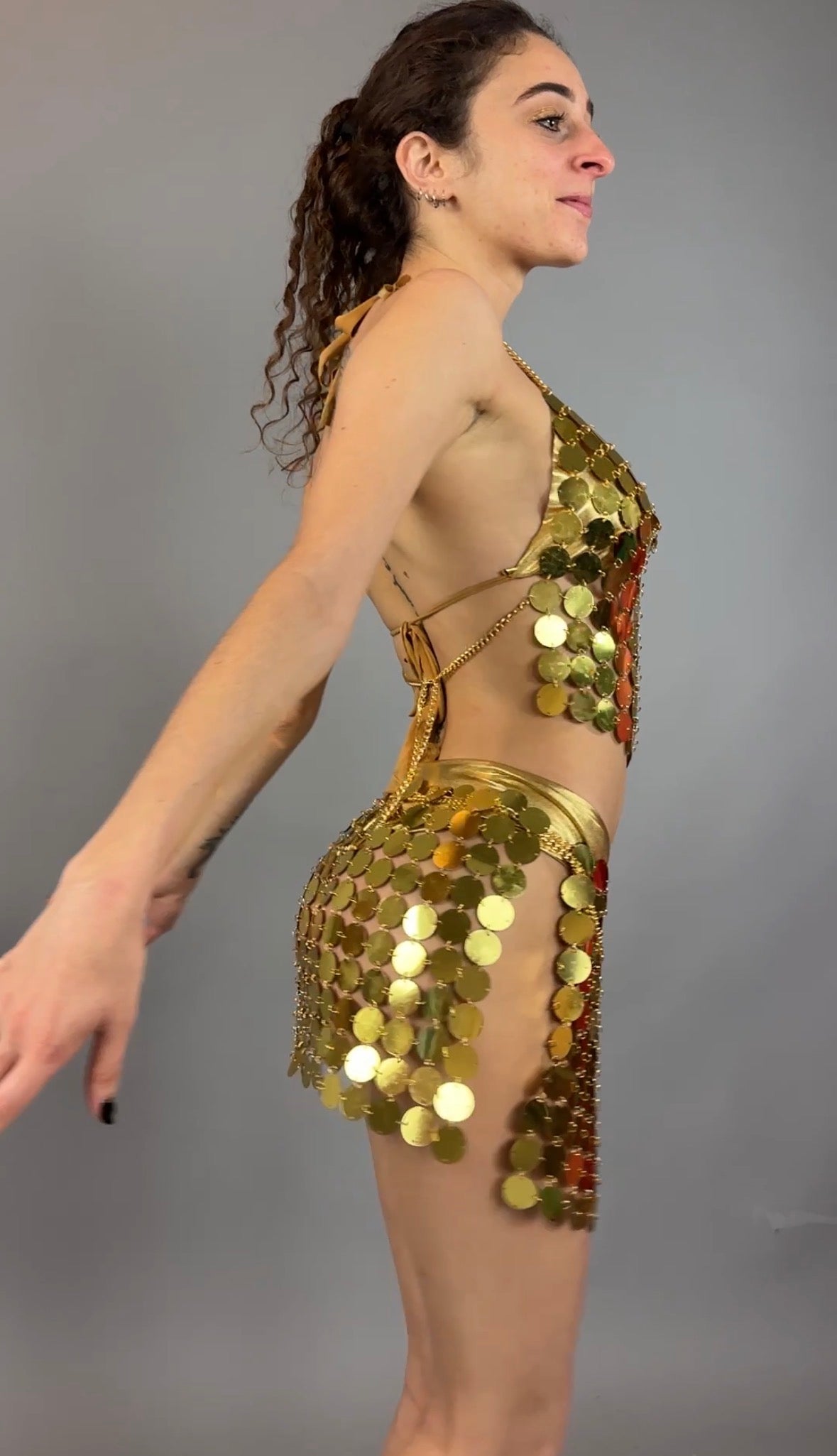 A woman poses sideways with arms outstretched in the Gold Chain Two-Piece Set for Performers costume, featuring a supportive crop top, against a gray background.
