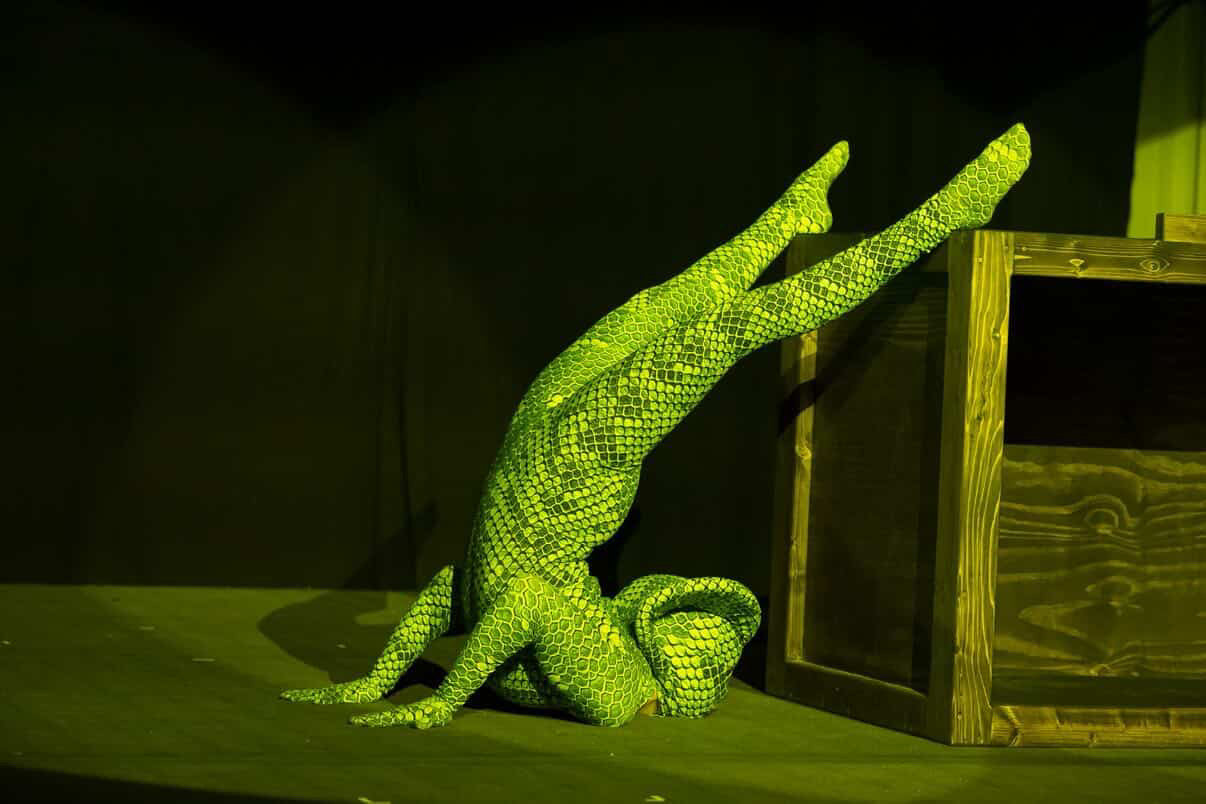 A performer in a Green pixel Cobra Costume Snake Print Catsuit balances on their shoulders with legs extended upward, displaying impressive contortionist skills and poise on stage.