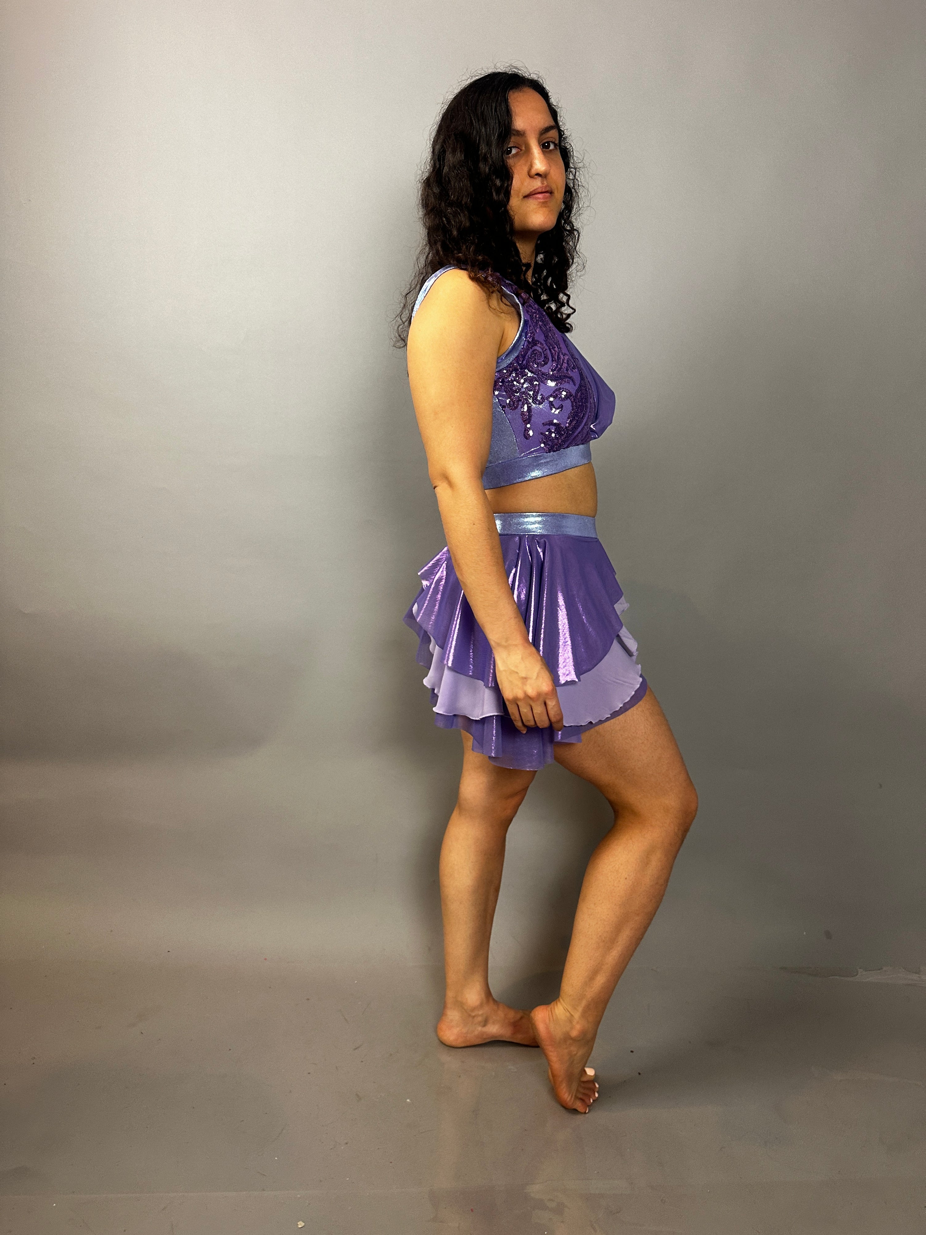 Woman with curly hair wears a Lavender Purple Sequin Dance Costume—Fire Dancer Outfit with high waist shorts, half skirt, and crop top in 4-way stretch mesh—standing barefoot against a plain gray background.