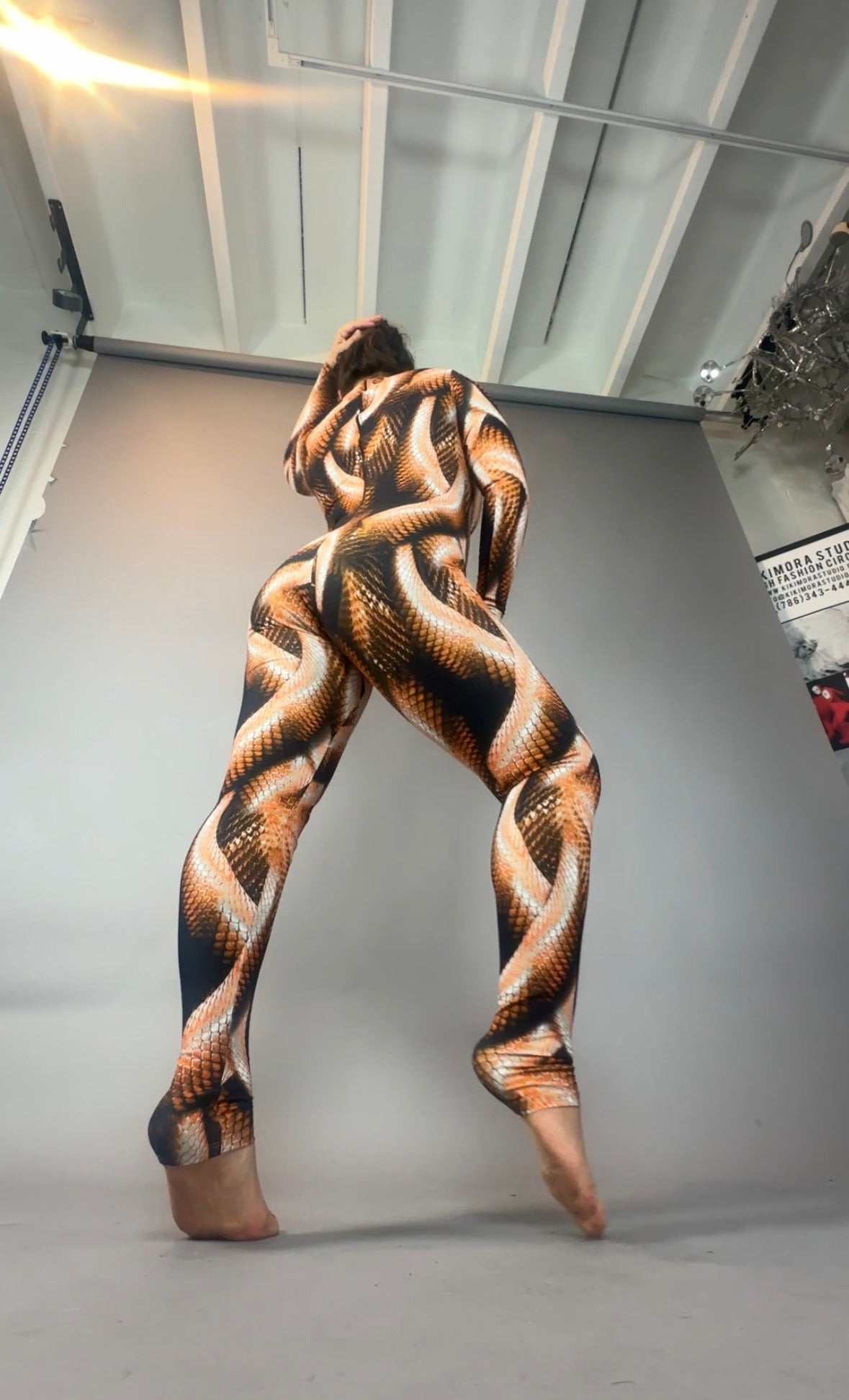 A person stands on tiptoes in a Brown Snake print Catsuit with turtleneck and long sleeves, posed against a plain studio backdrop.