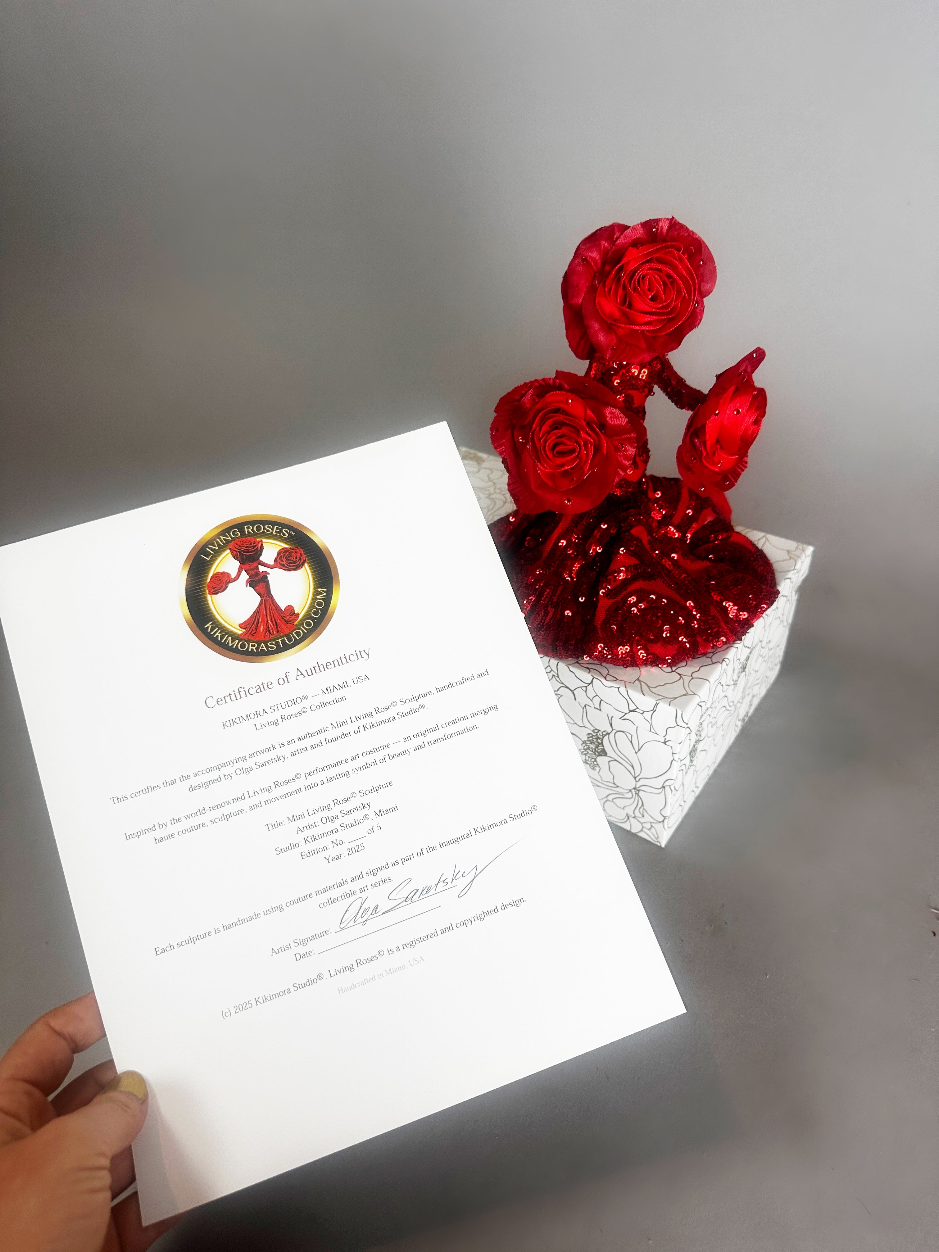 A hand holds a certificate beside a white box with three Mini Living Rose sculptures—doll ornaments by Kikimora Studio, each handcrafted and sparkling with red glitter.