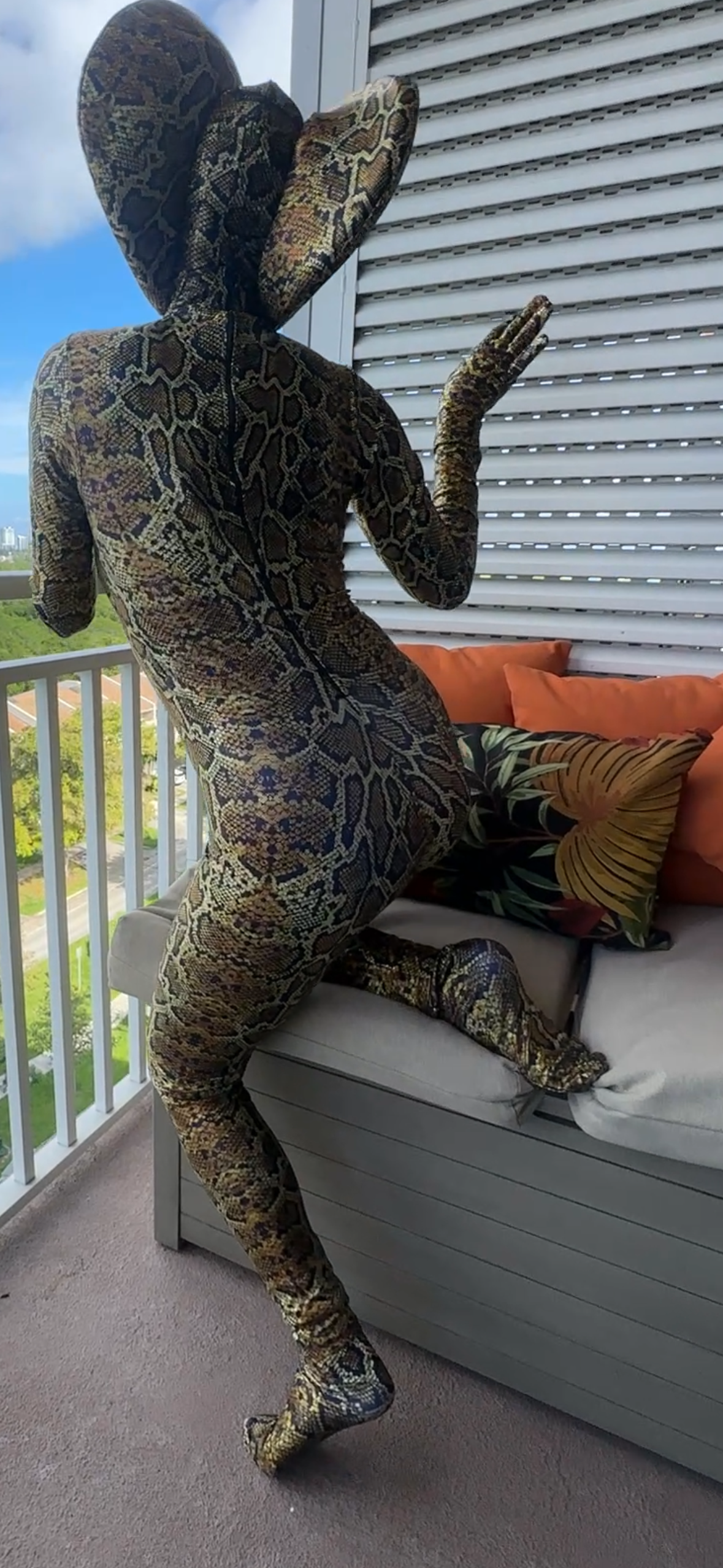 Wearing the Green Cobra Snake Costume, a contortionist poses on a balcony bench with orange and leaf-print pillows, completing the gymnastic outfit with a striking snake headpiece.