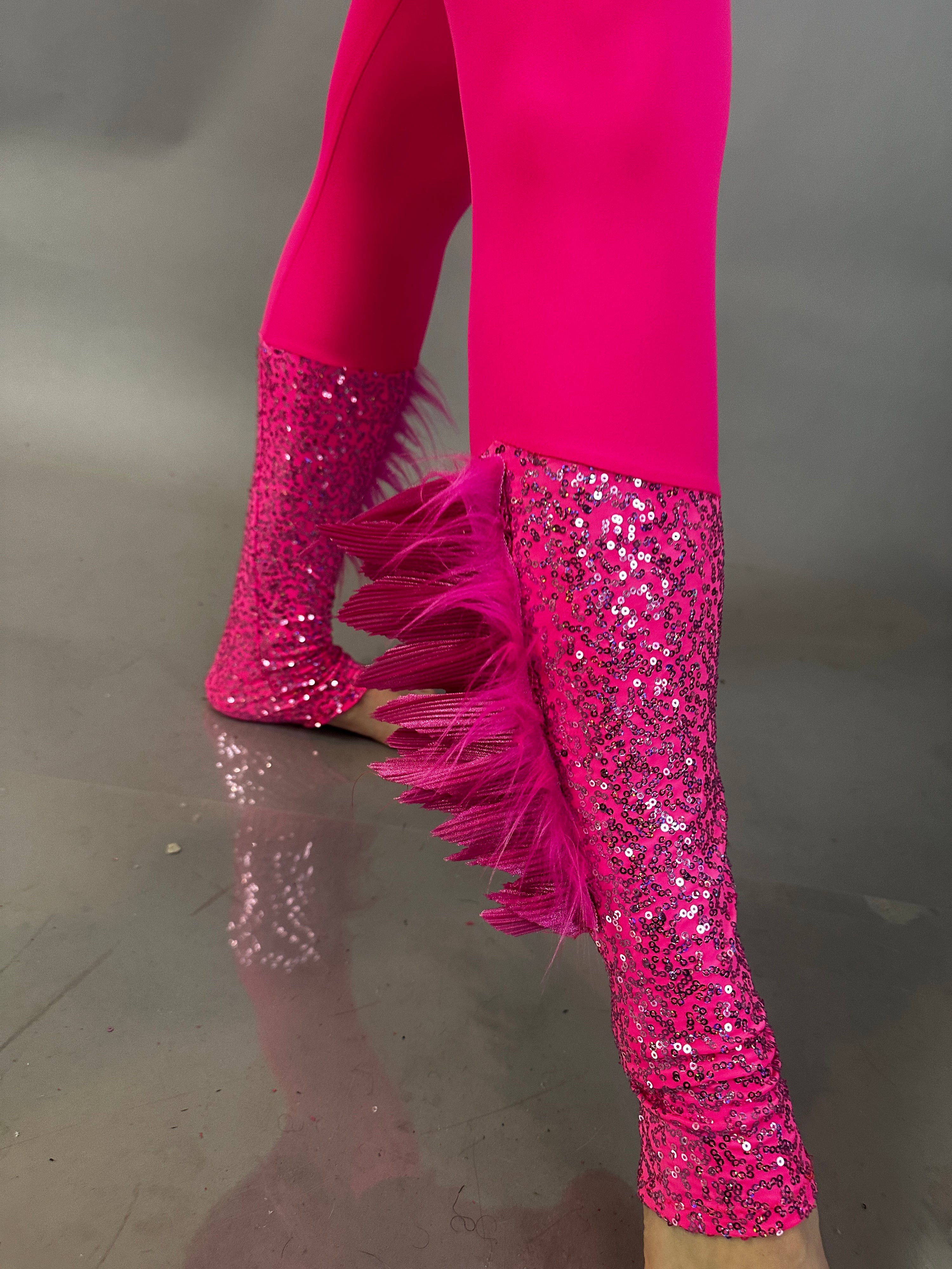 A person confidently wears the Pink Bird full costume for aerial and dancers, featuring bright pink sequin leggings with feather trim, standing on a reflective surface.