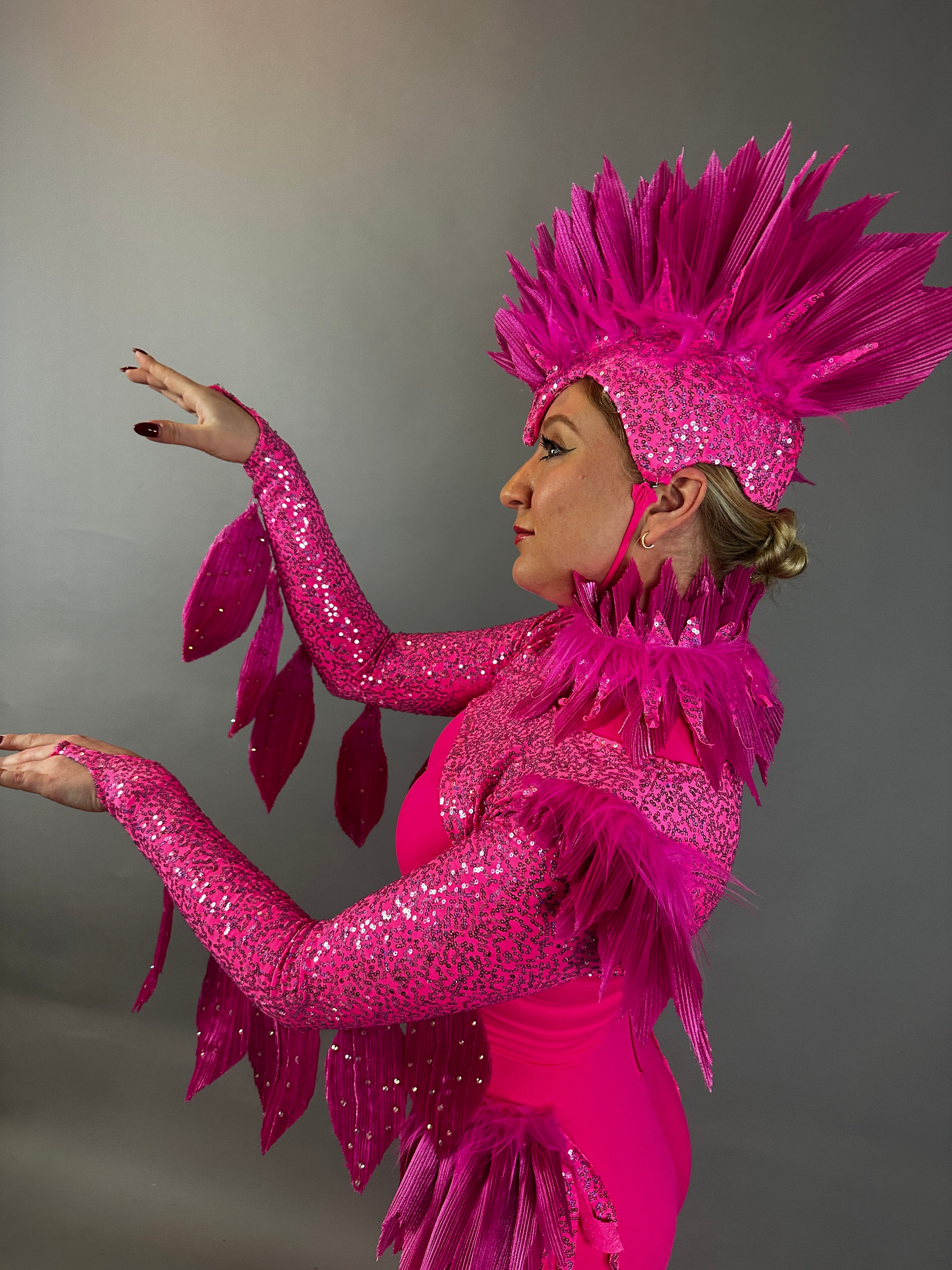 A person in profile wears the Pink bird full costume for aerial and dancers, featuring bright pink feathers and sequins, striking a dramatic pose with raised arms.