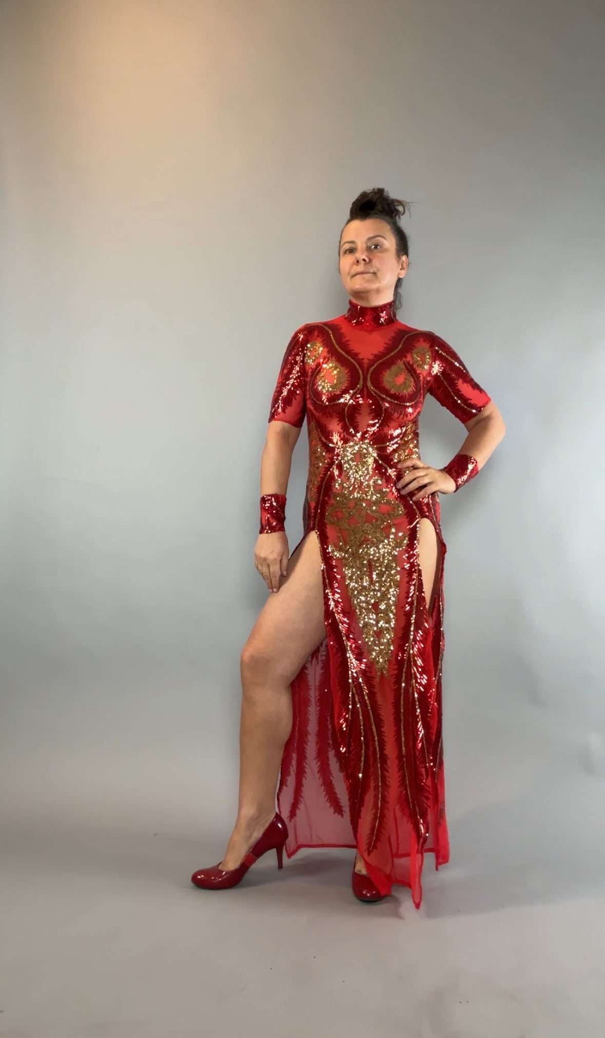 Woman in a Red and Gold Sequin Sheer Dress, sleeveless with hand accessories, poses confidently against a gray backdrop.