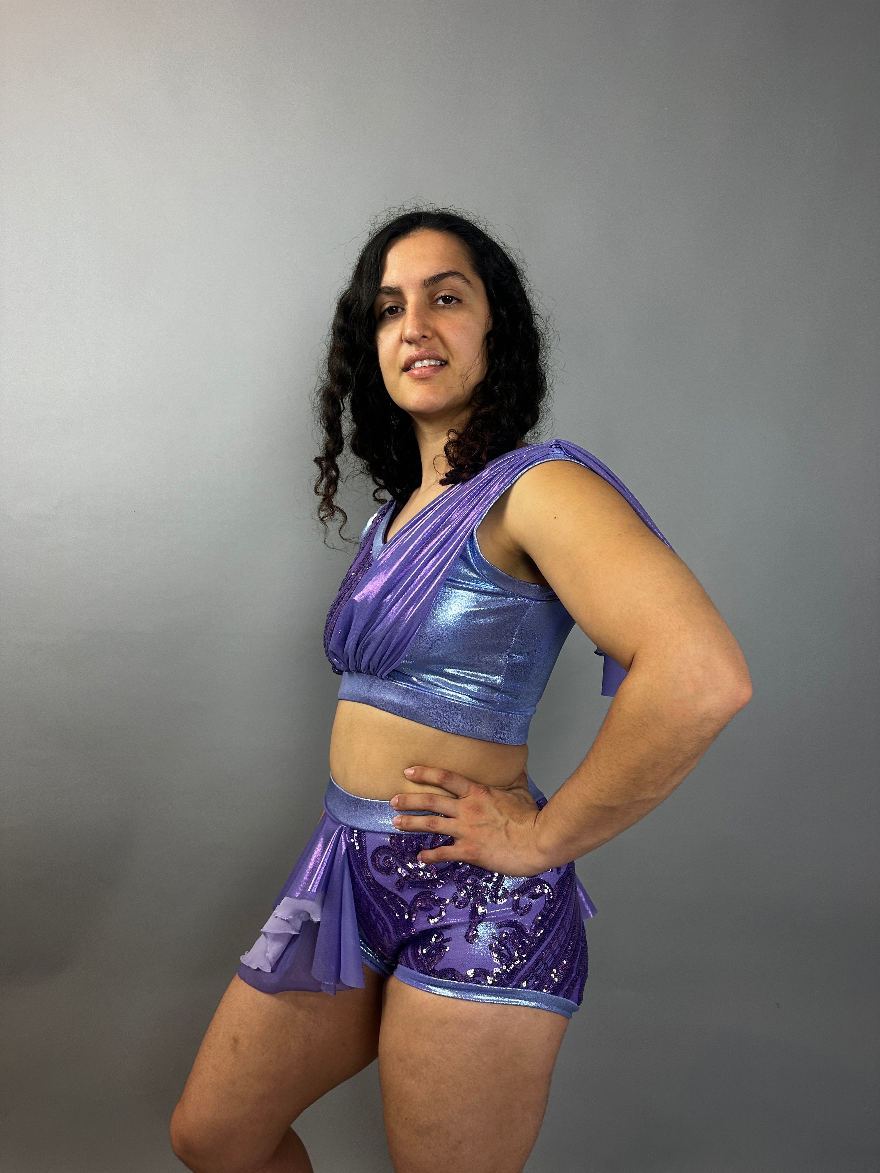 Wearing the Lavender Purple Sequin Dance Costume—featuring high waist shorts with a half skirt, crop top, and 4-way stretch mesh—the person smiles brightly against a plain gray background.