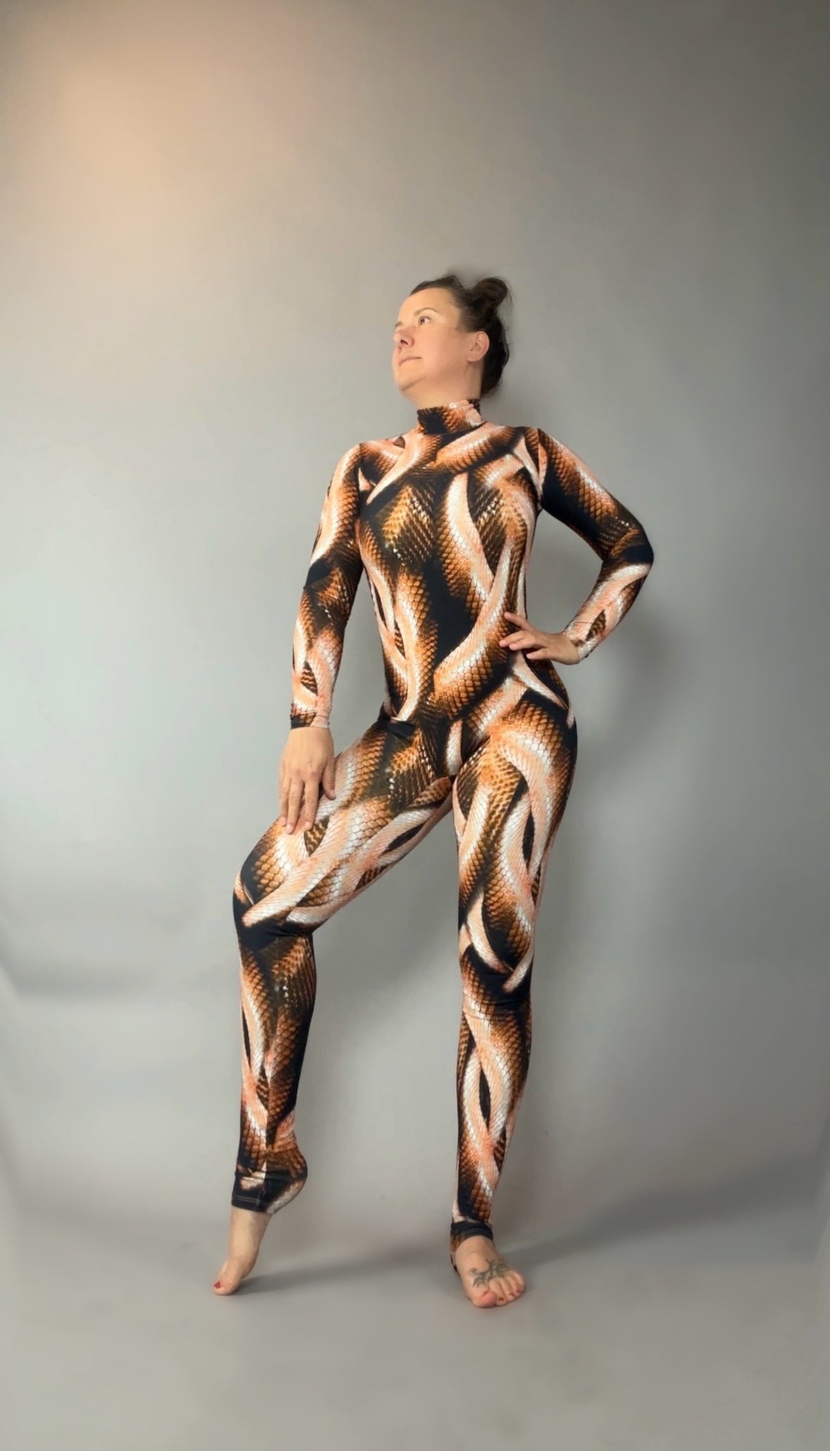 Person poses in a Brown Snake print Catsuit featuring a turtleneck and long sleeves against a plain gray background.