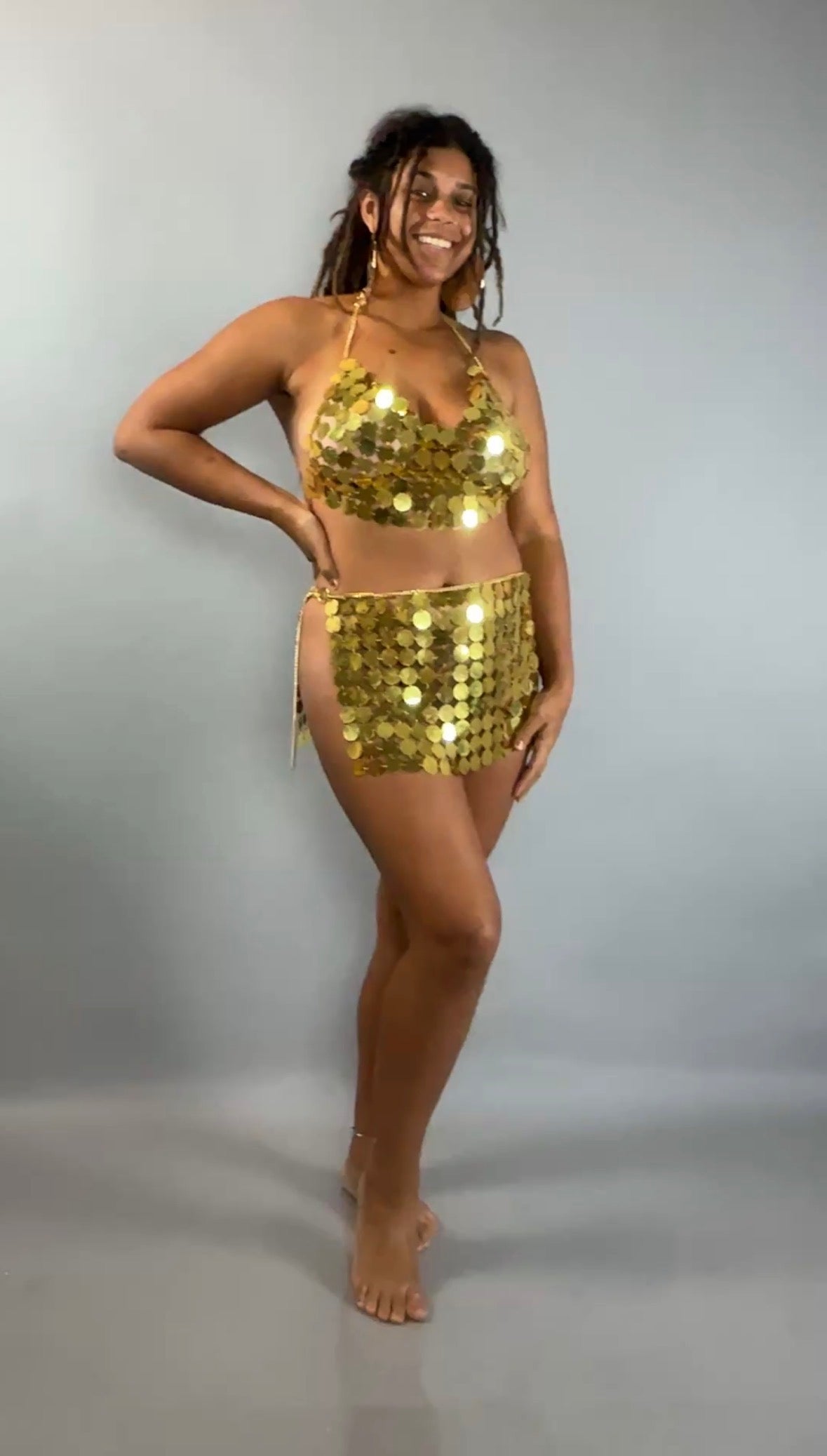 A smiling woman models the Gold Chain Two-Piece Set for Performers—featuring a supportive crop top and skirt—against a plain gray background.