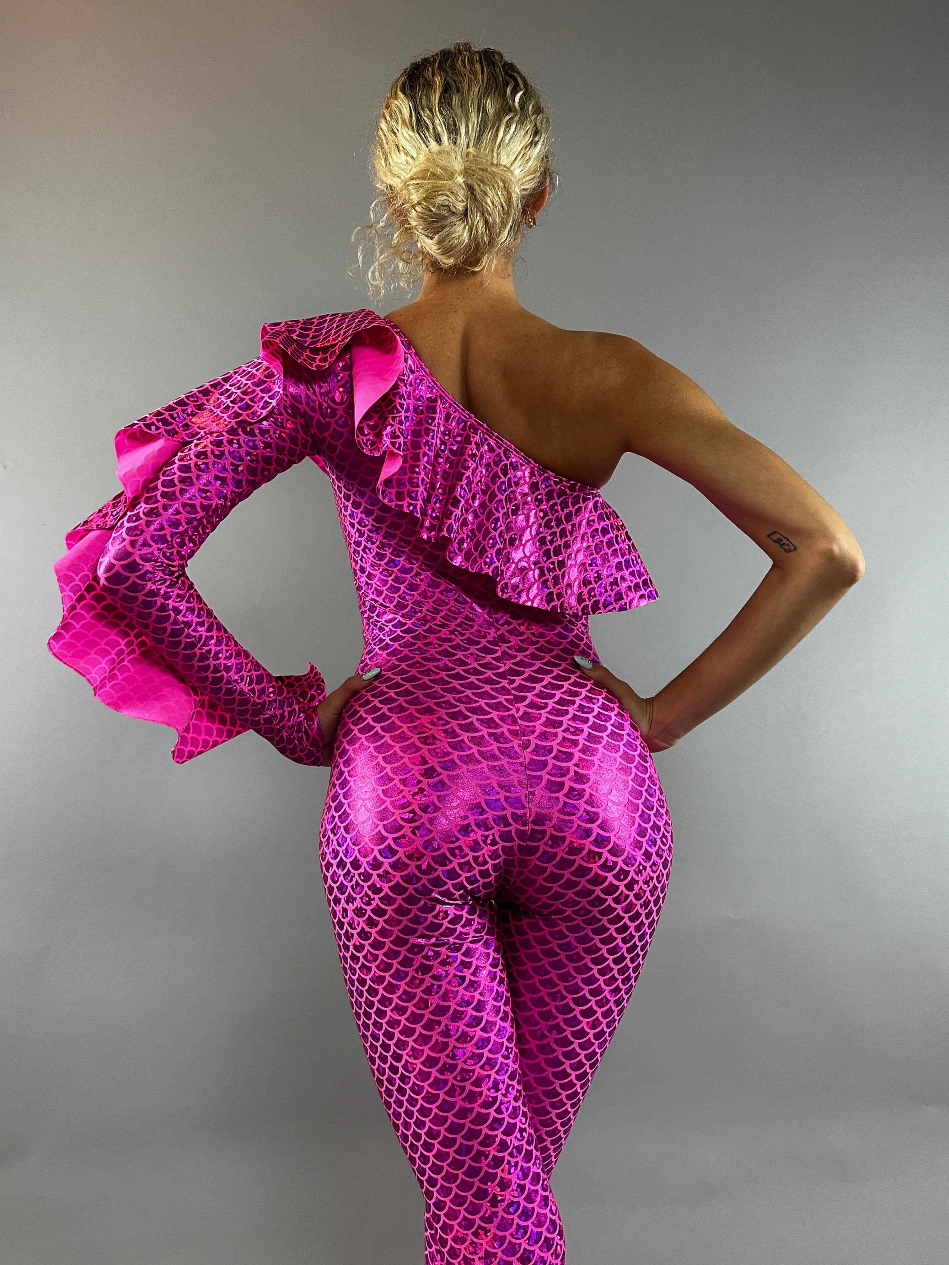 A blonde-haired person in a bun wears the Ruffle Catsuit—an off-shoulder, asymmetrical spandex jumpsuit in shiny pink—posing against a gray backdrop.