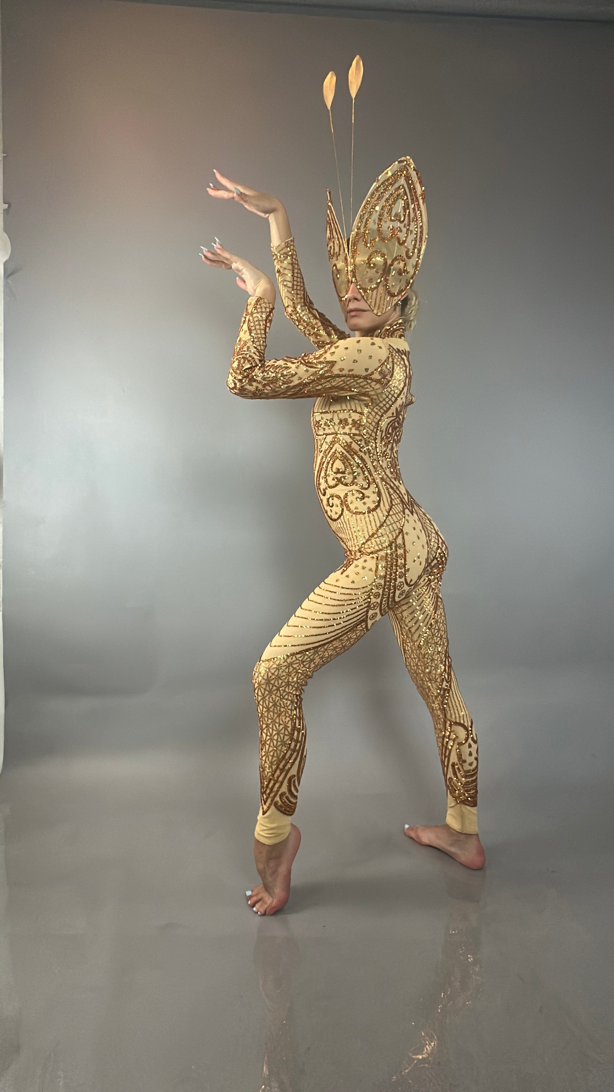 Wearing the Black Sequin Butterfly Headpiece with feather antennae, a model poses elegantly on tiptoe in a gold ornate bodysuit against a plain gray background.