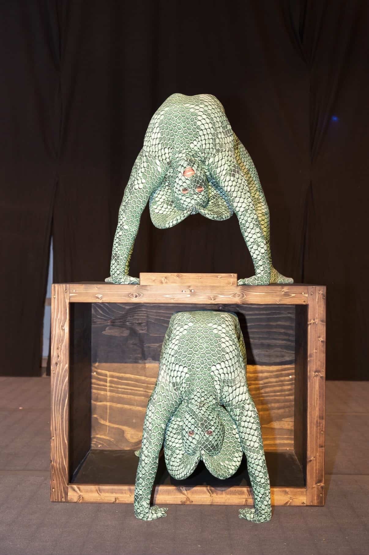 Two performers in Green Pixel Cobra Costumes—snake print catsuits with headdress—pose on and inside a wooden box, their serpent cosplay creating a striking visual against the dark background.