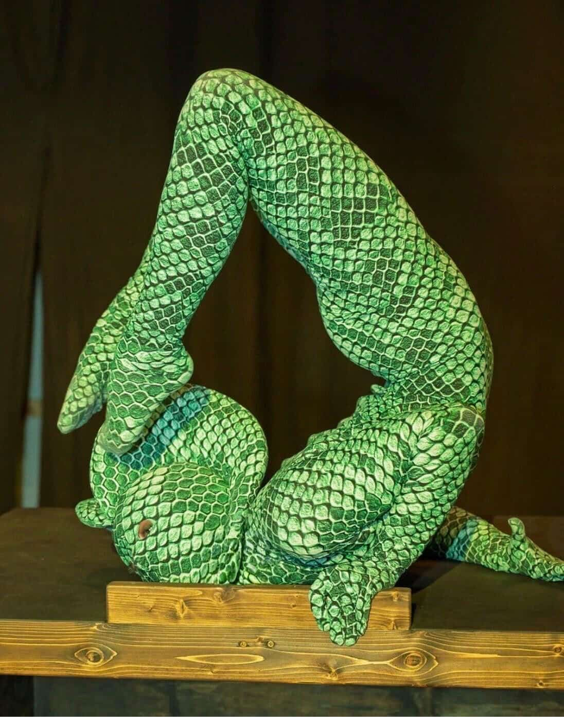 Wearing the Green Pixel Cobra Costume—snake print catsuit with headdress—a person performs a flexible backbend on a wooden surface.