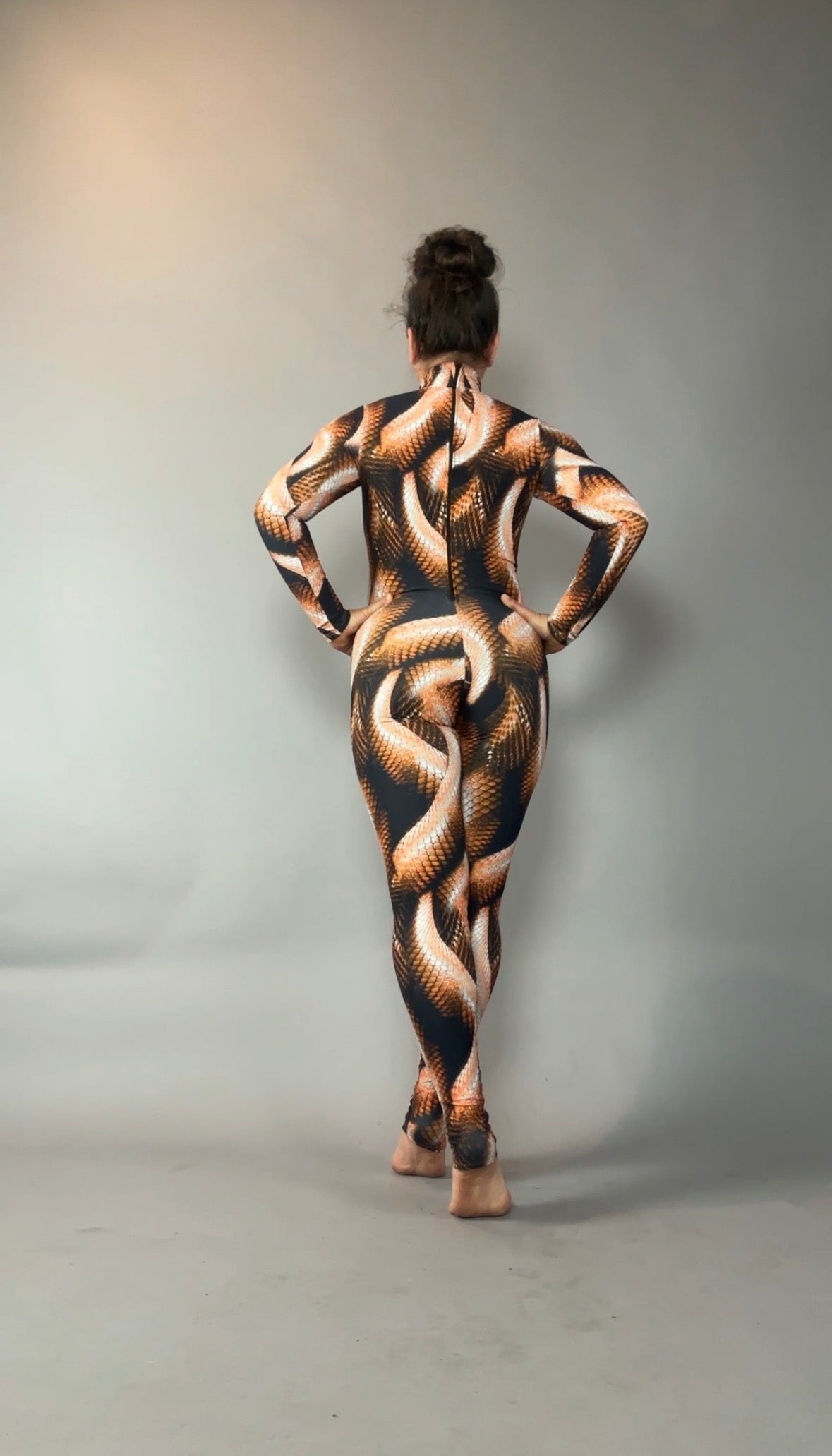 A person stands barefoot in a brown snake print catsuit with turtleneck and long sleeves, facing away against a plain gray background.