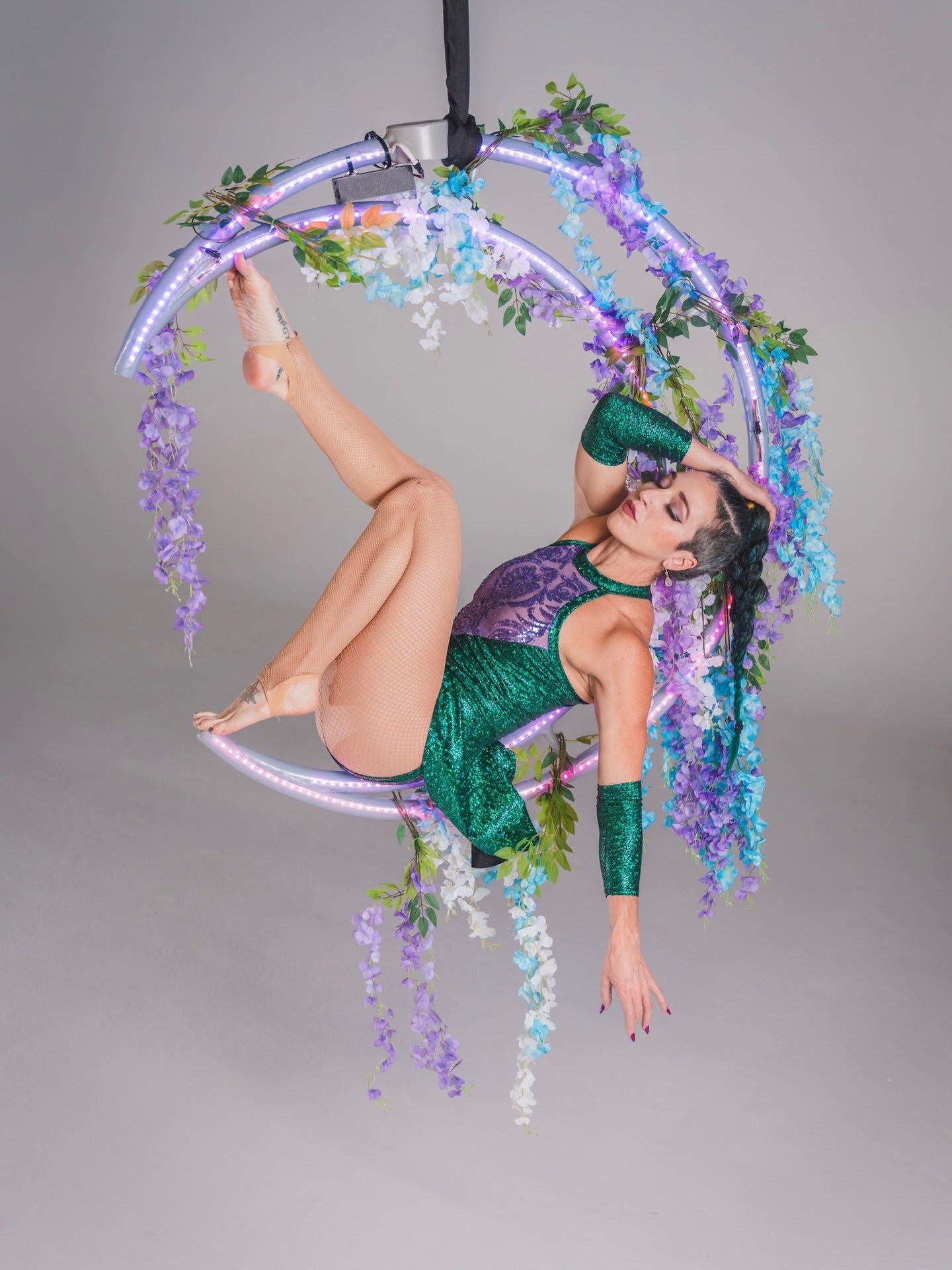 Aerial performer poses on a floral LED-lit hoop, suspended in the air against a gray background, wearing the Shattered Emerald Skater Leotard with halter neck and sheer sequin details.