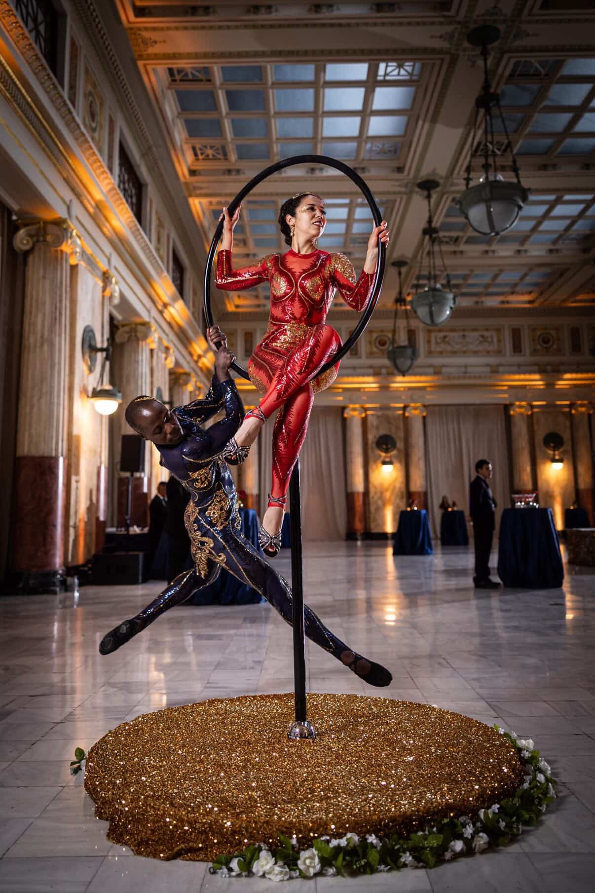 Two acrobats perform on a hoop in an elegant hall, wearing custom-made Showgirl Costumes: black sequin bodysuits with sheer details, designed for both women and men.
