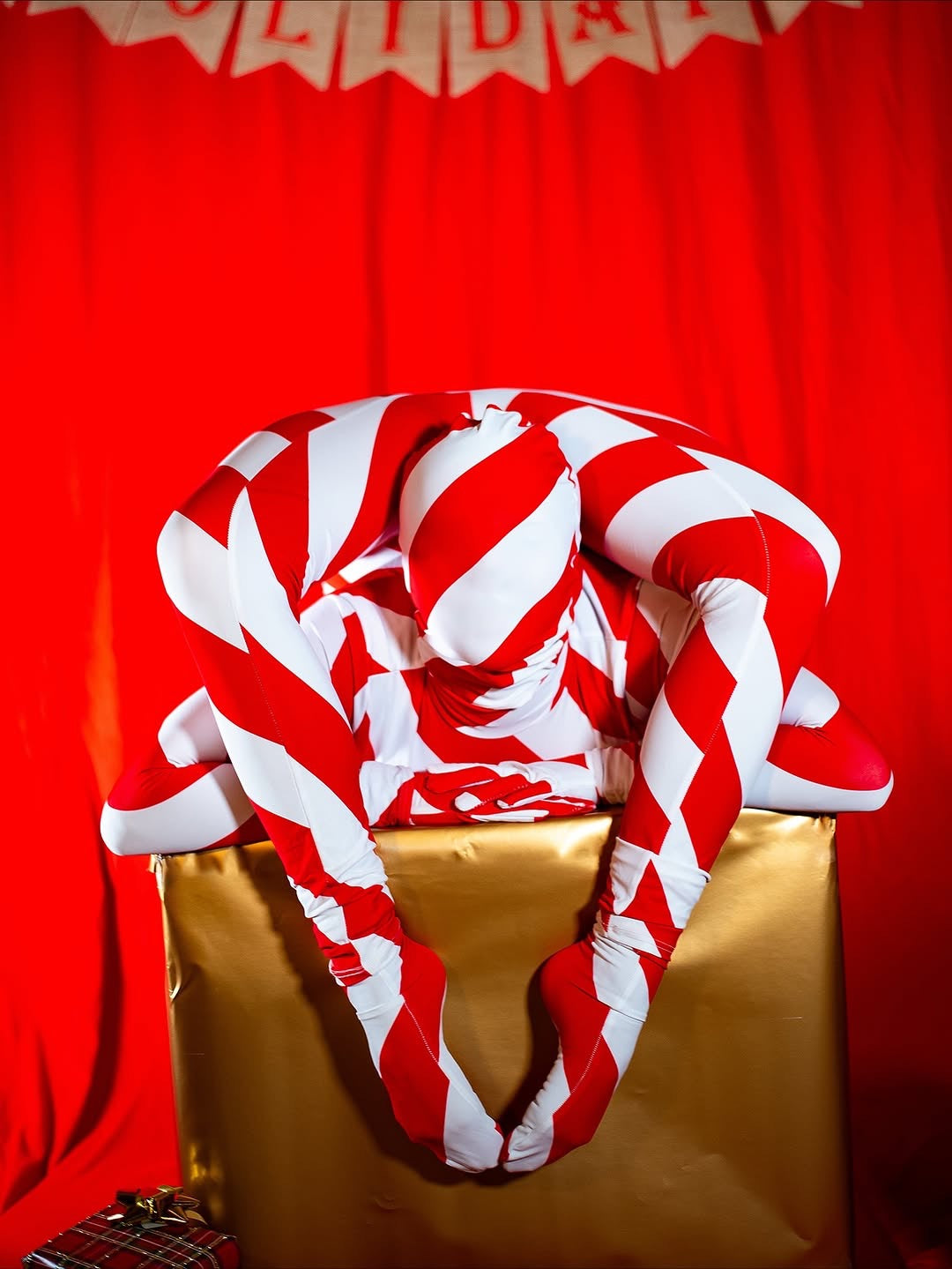 A model wearing the Candy Cane Bodysuit with a turtleneck and full sleeves poses on a gold box against a red backdrop.