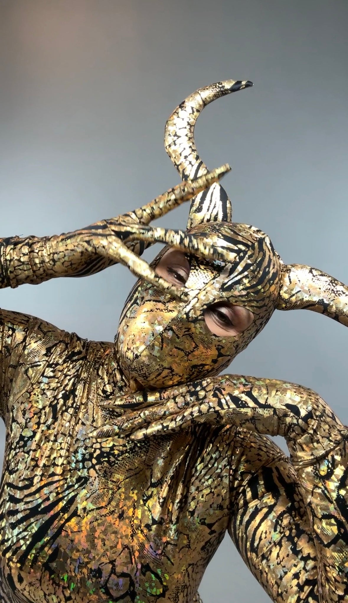 A person wearing a Gold & Black Gazelle Creature Costume, which includes a full sleeve catsuit, gloves, socks, and an elaborate horned headdress, strikes a dramatic pose with face paint and hands framing their face.