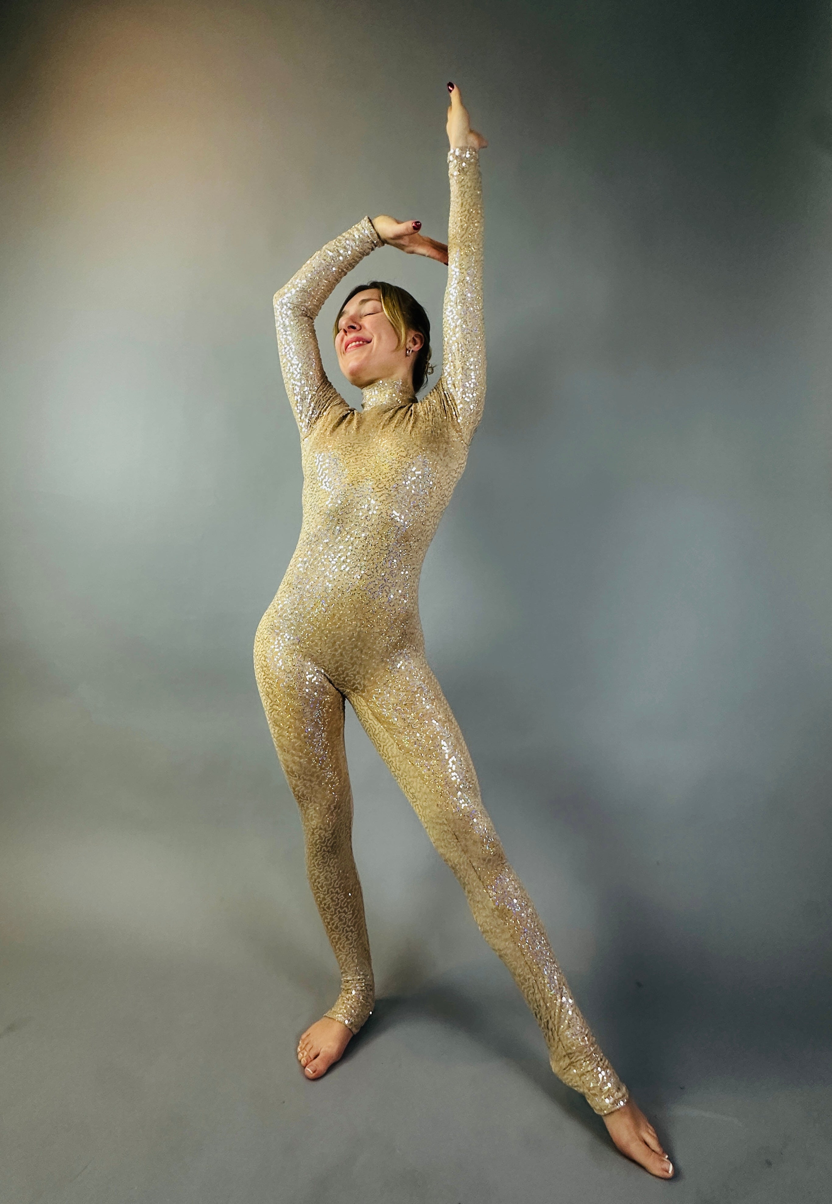 Barefoot and radiant, a model showcases the Champagne Fairy Sequin Catsuit with open back, turtleneck, and sleeves, posing with one arm raised and sparkling against a gray background.