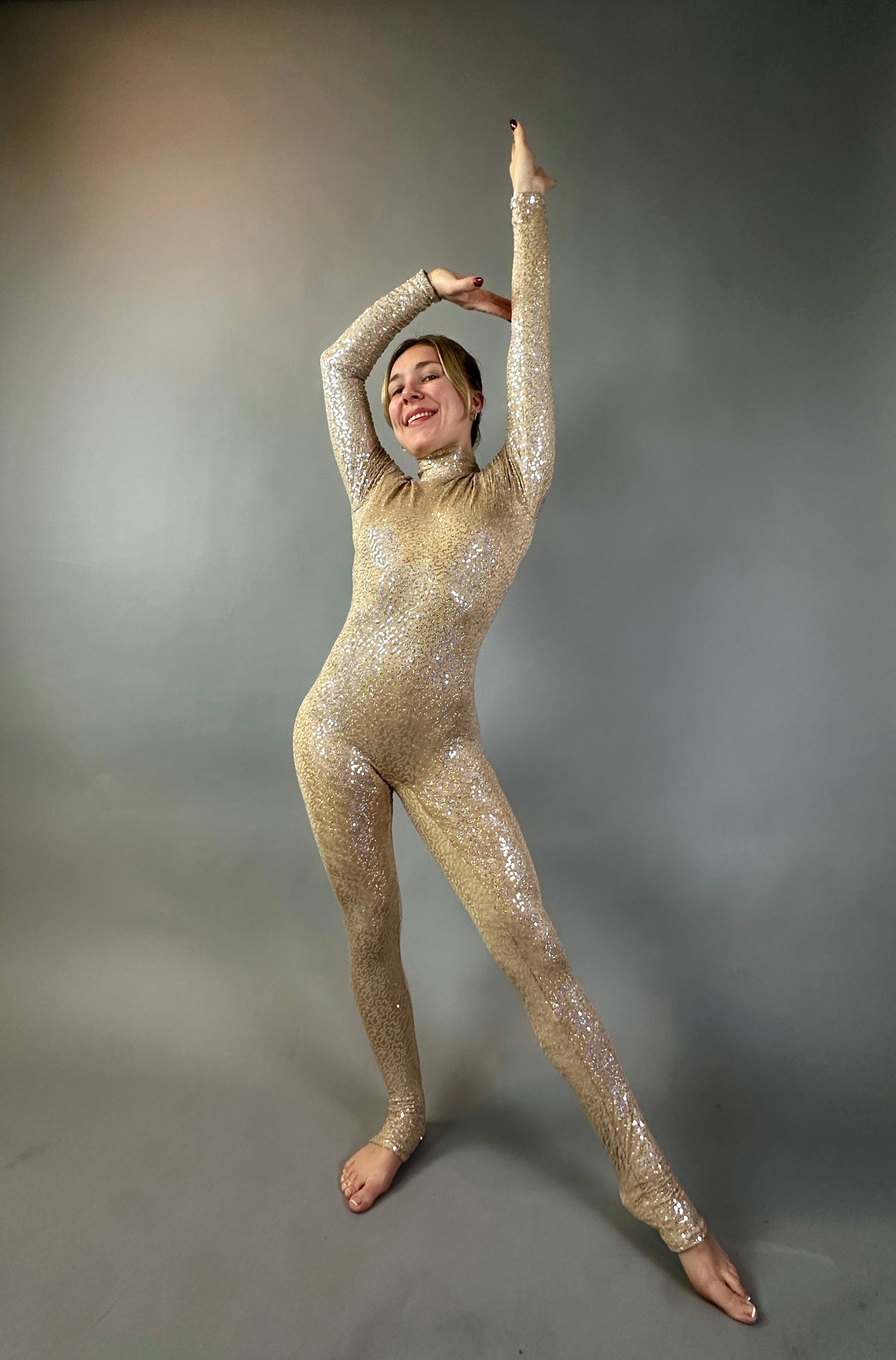 Model poses joyfully with arms raised in the Champagne Fairy Sequin Catsuit, featuring an open back, turtleneck, and sleeves, against a plain grey background.