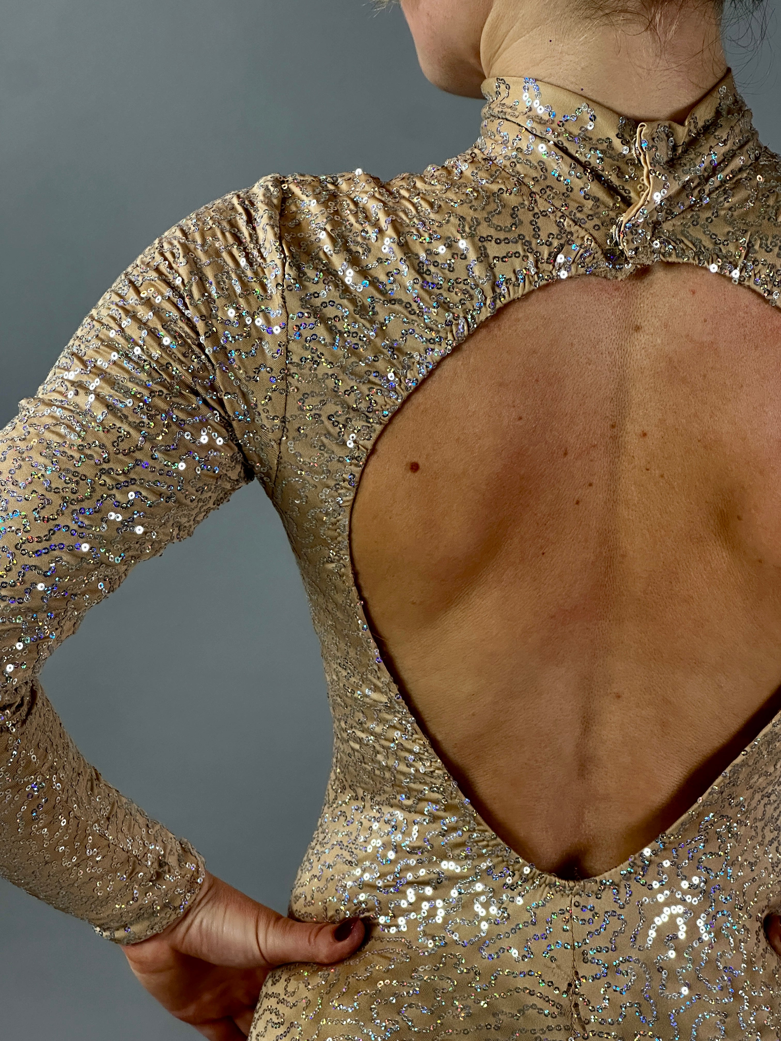 A person wearing the Champagne Fairy Sequin Catsuit—featuring a turtleneck, sleeves, and an open back—sparkles against a gray background, evoking glamorous performance style.