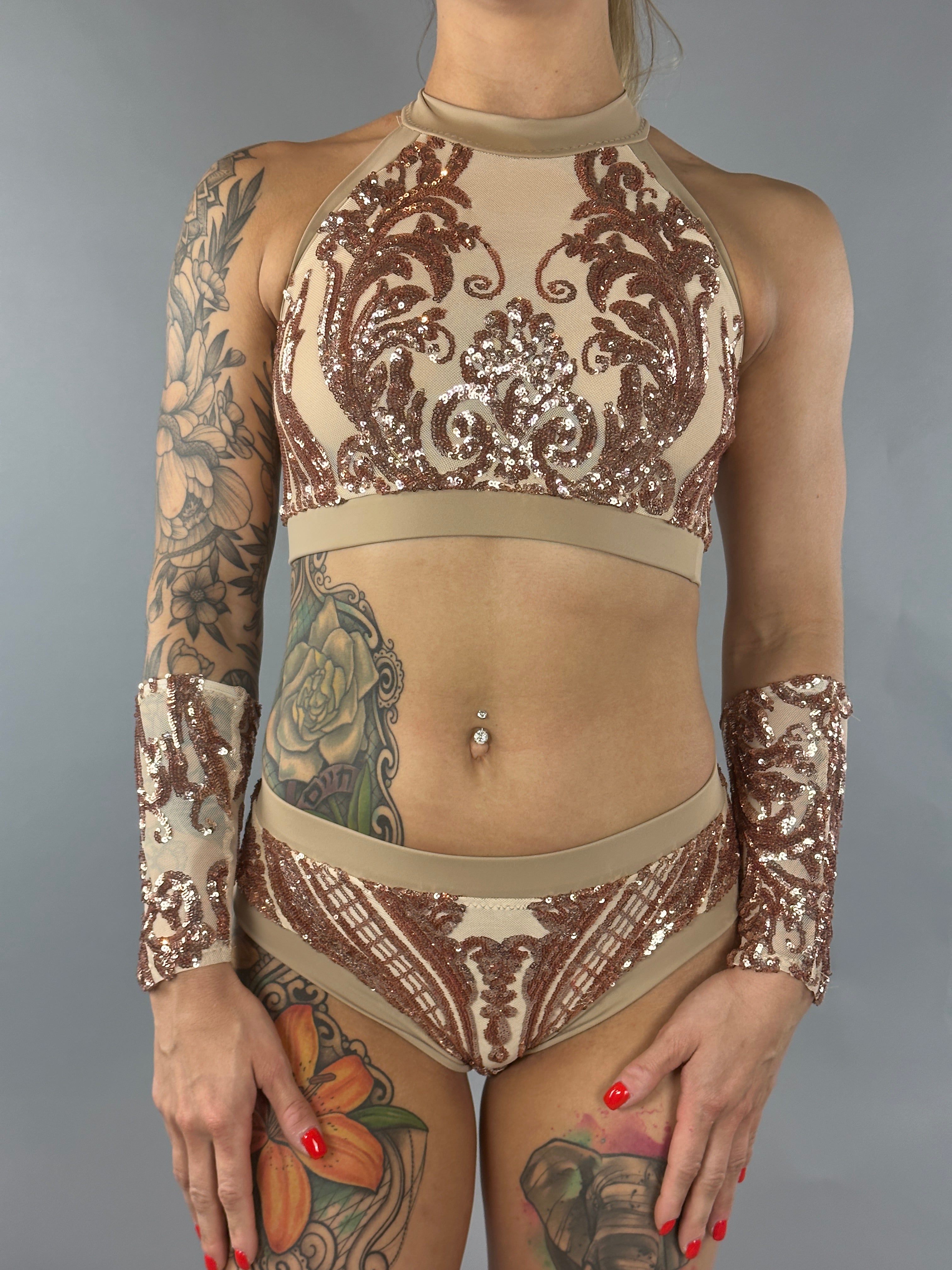 A person wearing a decorative Sequin Crop Top and Bottom Set with a halter neck, in beige and brown embellished with intricate designs and shimmering gold sequins, complete with matching handcuffs. This dazzling ensemble mirrors the elegance of high-end performance wear.