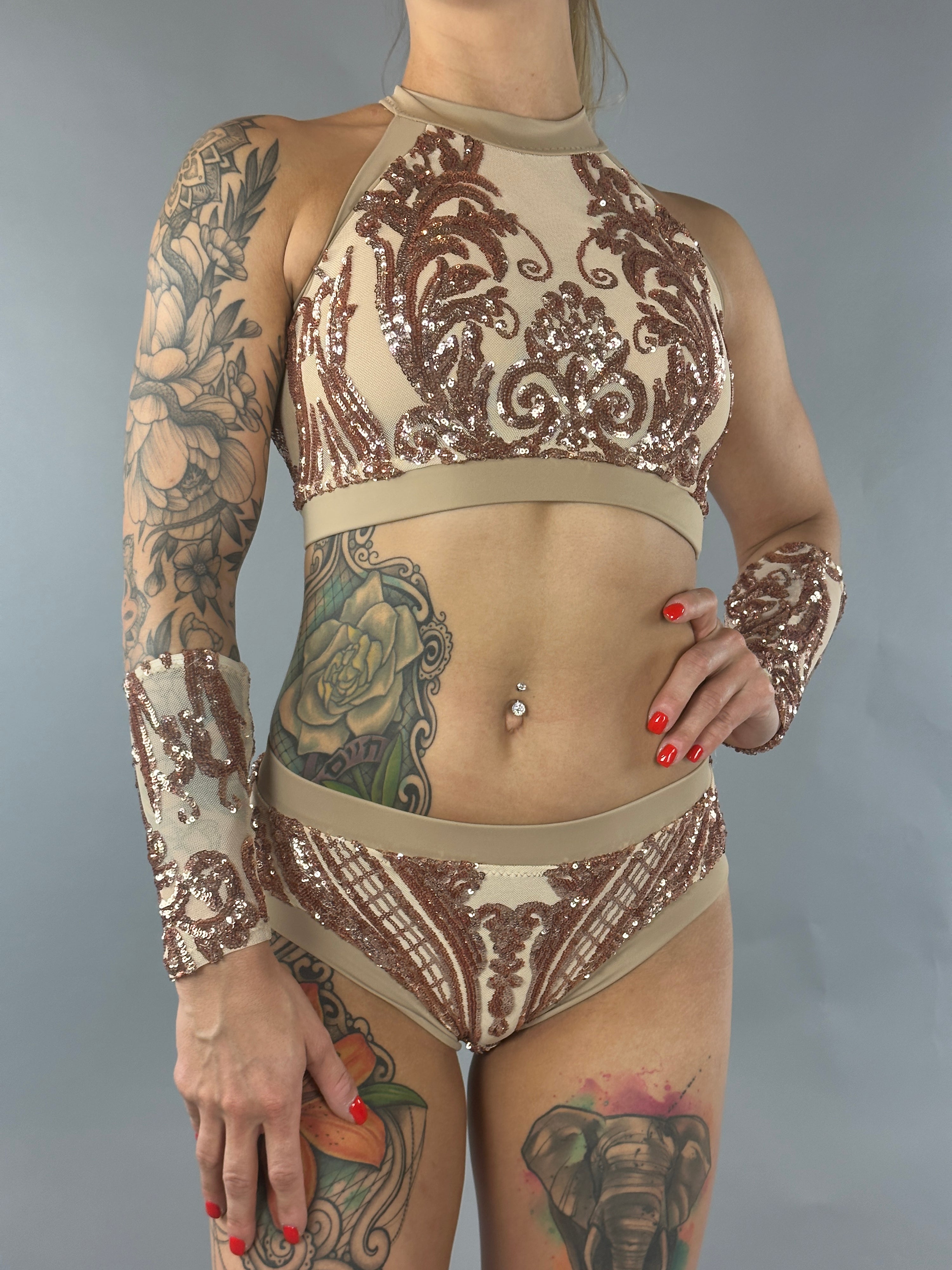 A person in a Sequin Crop Top and Bottom Set with floral tattoos and red nails stands against a gray background, shimmering gold sequins catching the light.