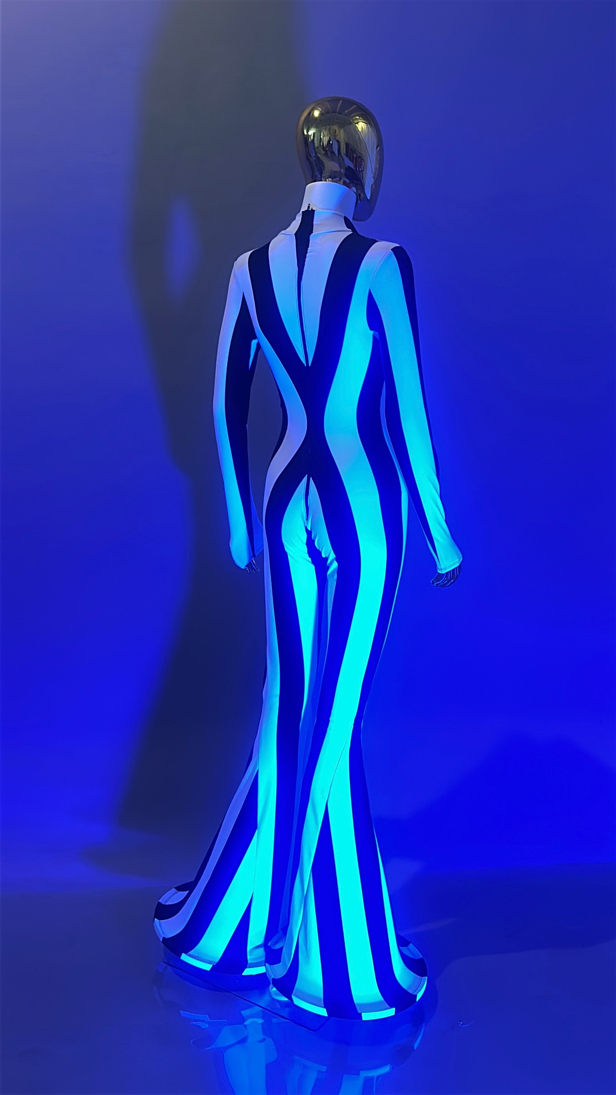 A mannequin wears the Rave White Striped Catsuit with bellbottom legs and a gold helmet, standing before a blue-lit backdrop for a bold festival performer vibe.