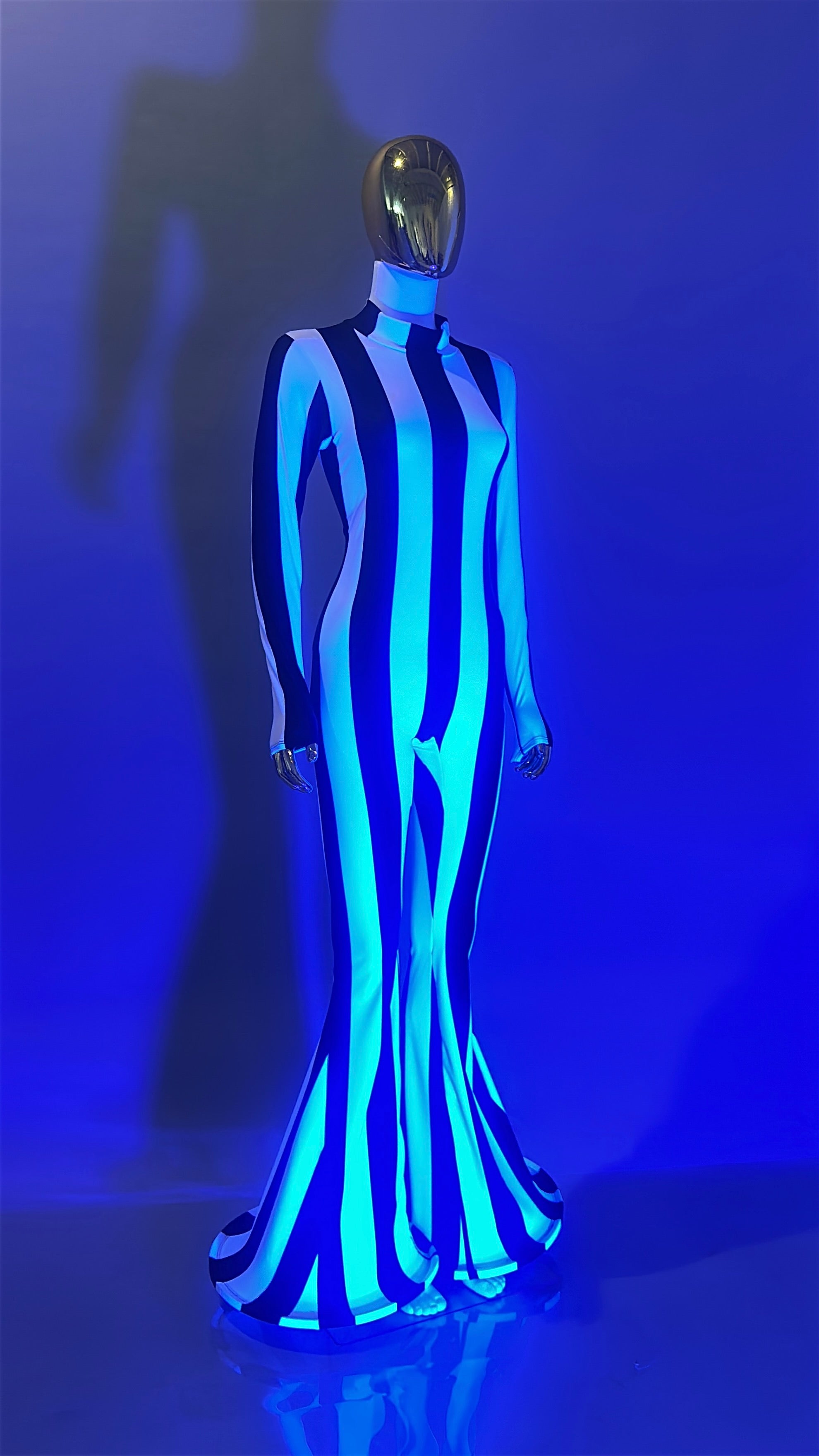 A mannequin wears the Rave White Striped Catsuit with bellbottom legs, featuring bold vertical stripes, set against a vivid blue background with dramatic lighting for a festival-ready look.