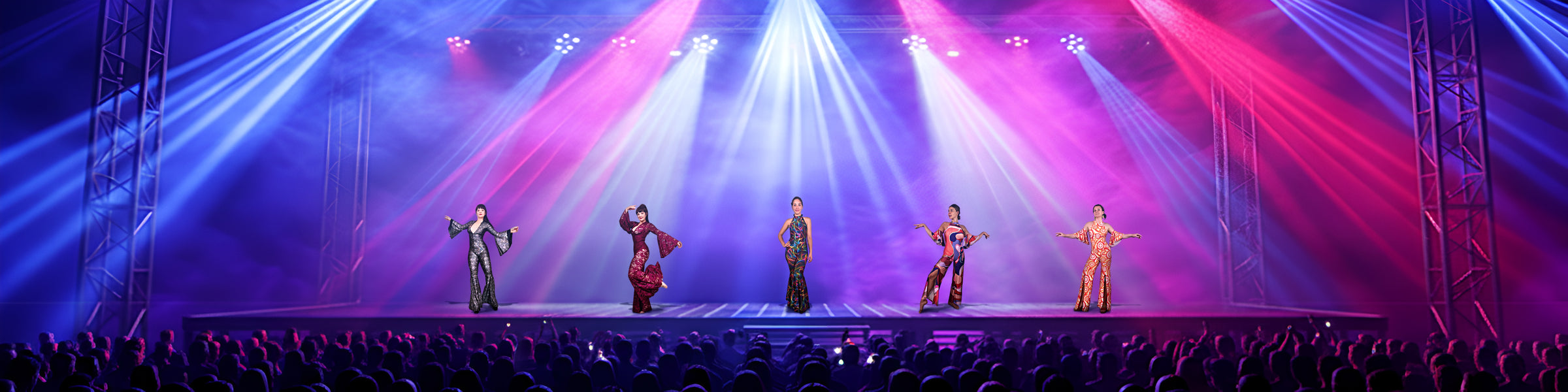 Five performers in colorful outfits pose on a brightly lit stage with an audience watching.