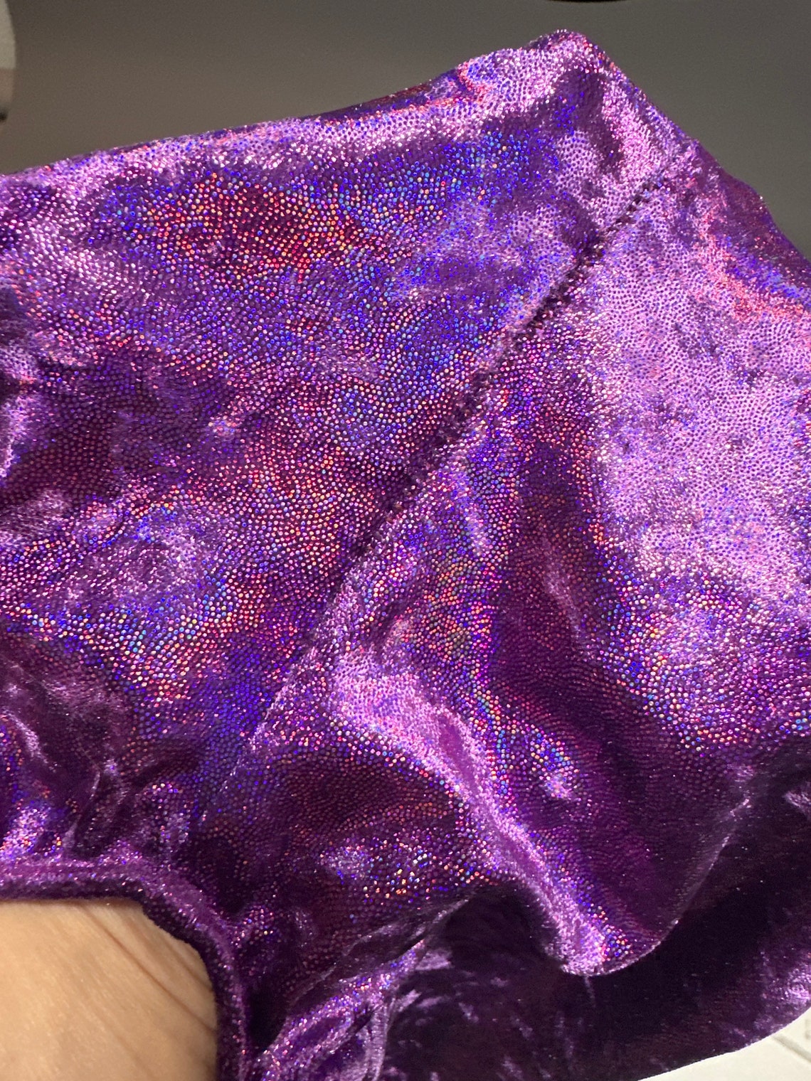 The Velvet Open Back Jumpsuit features shiny purple velvet with a reflective, iridescent texture and visible seams—ideal as exotic dance wear or a stunning festival outfit. Trending now for bold costume statements.