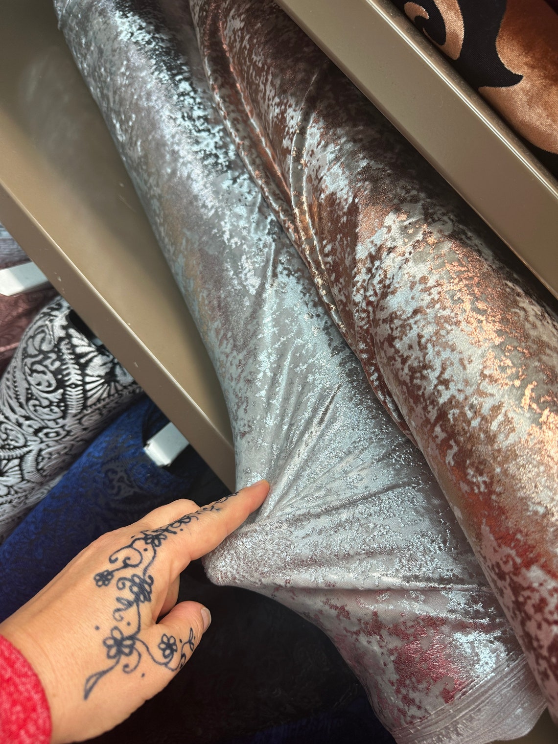 A hand with henna touches shiny silver and rose gold velvet rolls—perfect for crafting the Velvet Open Back Jumpsuit, Exotic Dance Wear, a trending festival outfit—displayed on a store shelf.