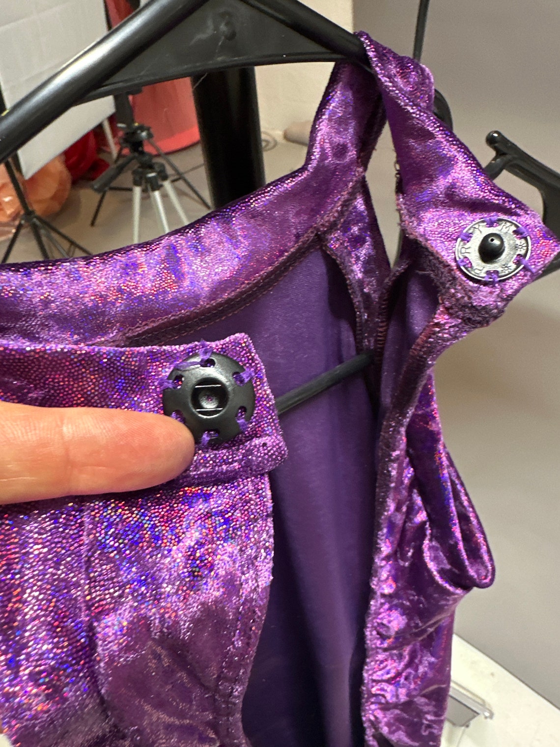 A hand holds a Velvet Open Back Jumpsuit, Exotic Dance Wear, Trending Now festival Outfit in sparkly purple with black snap buttons on the straps, hanging on a black hanger.