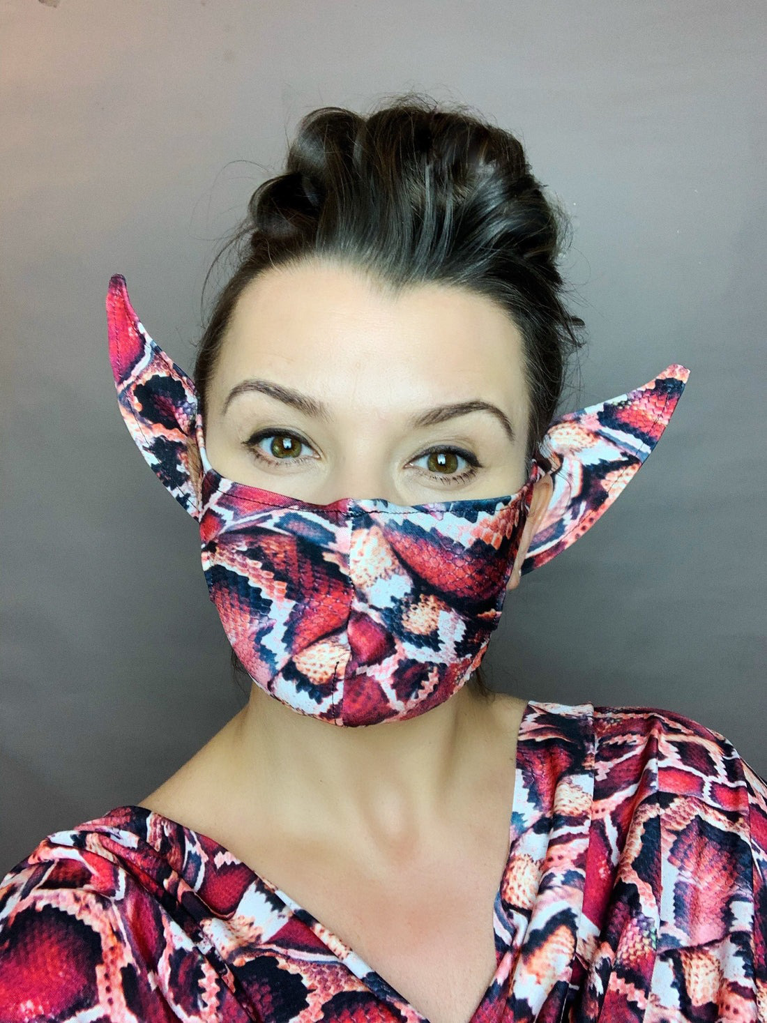 A woman poses in front of a plain background wearing the Elf Ears Face Mask for All Ages in snake print, styled as a reusable Mandalorian Baby Yoda costume, paired with a matching top.