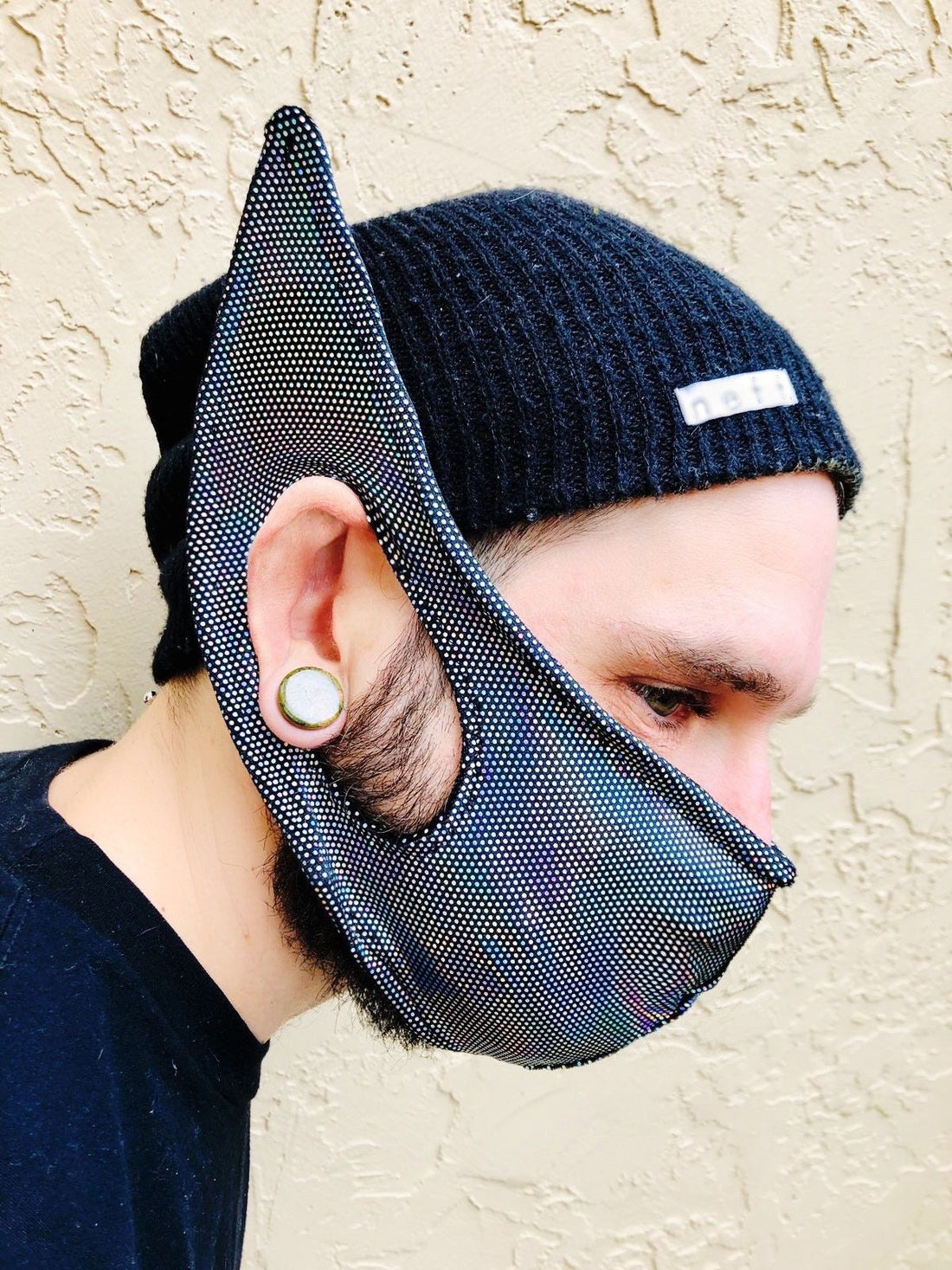 Person wearing the Elf Ears Face Mask for All Ages—funny mask inspired by Mandalorian Baby Yoda, a unique gift for men or women. Made in USA and styled with a black beanie and ear gauge for a standout look against a beige wall.