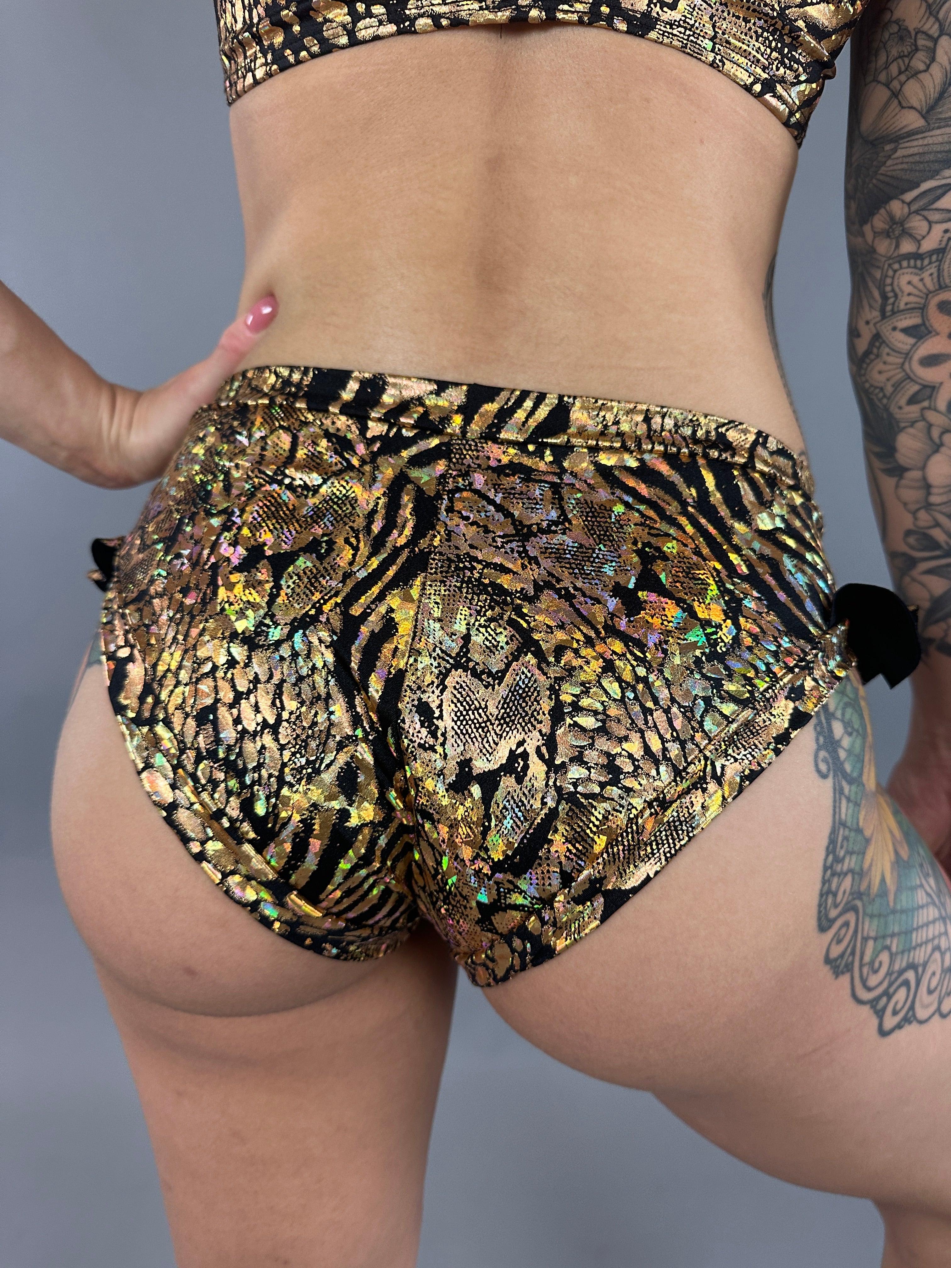 A person in Gold & Black Snakeskin Crop Top & Bottom Set, showcasing tiger and snake print shorts, stands against a gray background, highlighting their tattooed arm and leg.