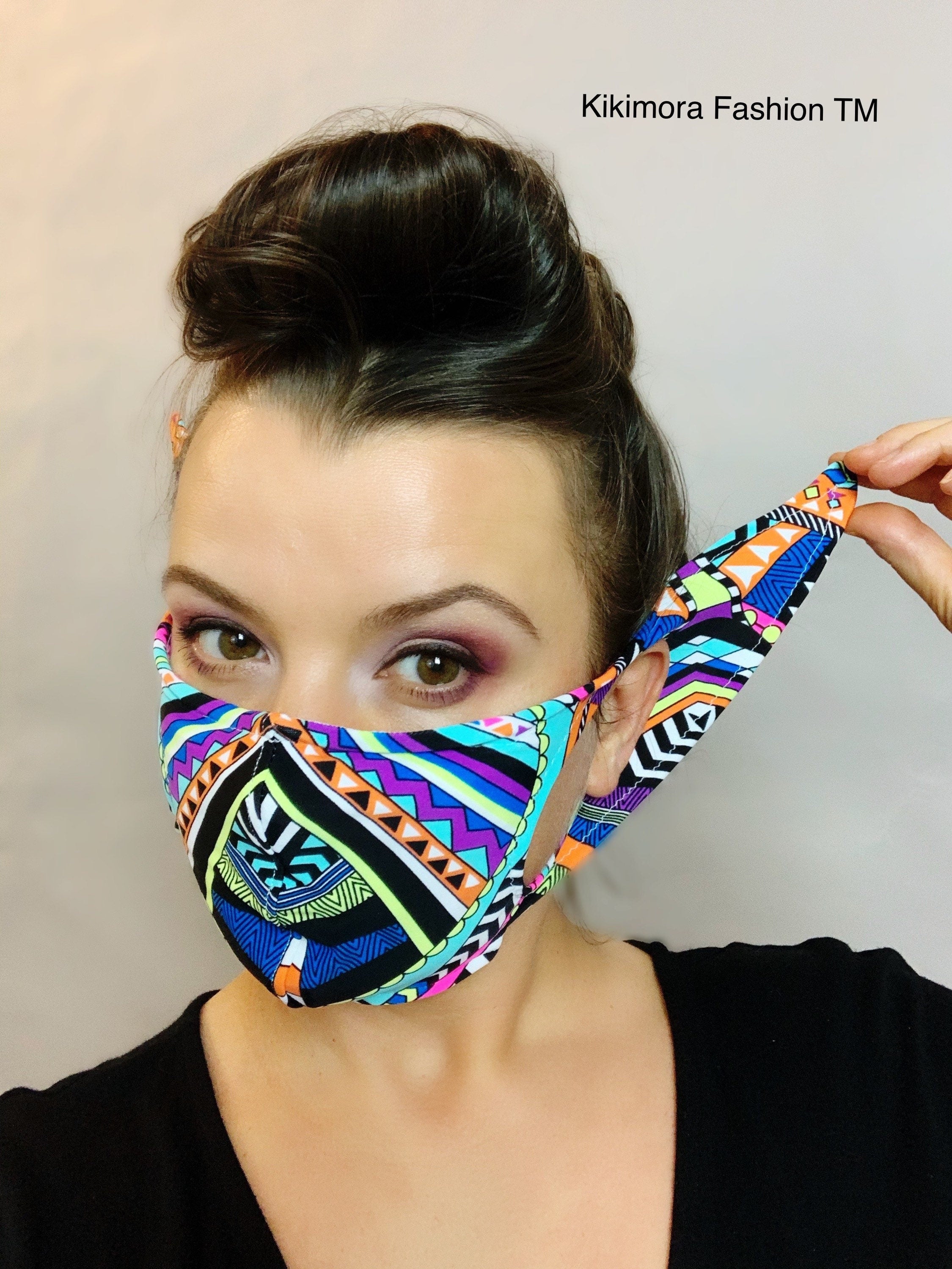 Woman wearing a colorful Glow Face Mask With 5 Layer HEPA Filter, pulling one strap, with "Kikimora Fashion TM" text in the cornerâhighlighting sustainable fashion with style.