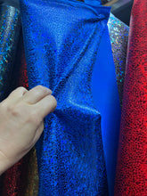A hand holds a blue, cracked-patterned fabric resembling the Exotic Dancewear jumpsuit, reminiscent of an aerialist costume and surrounded by colorful fabrics.