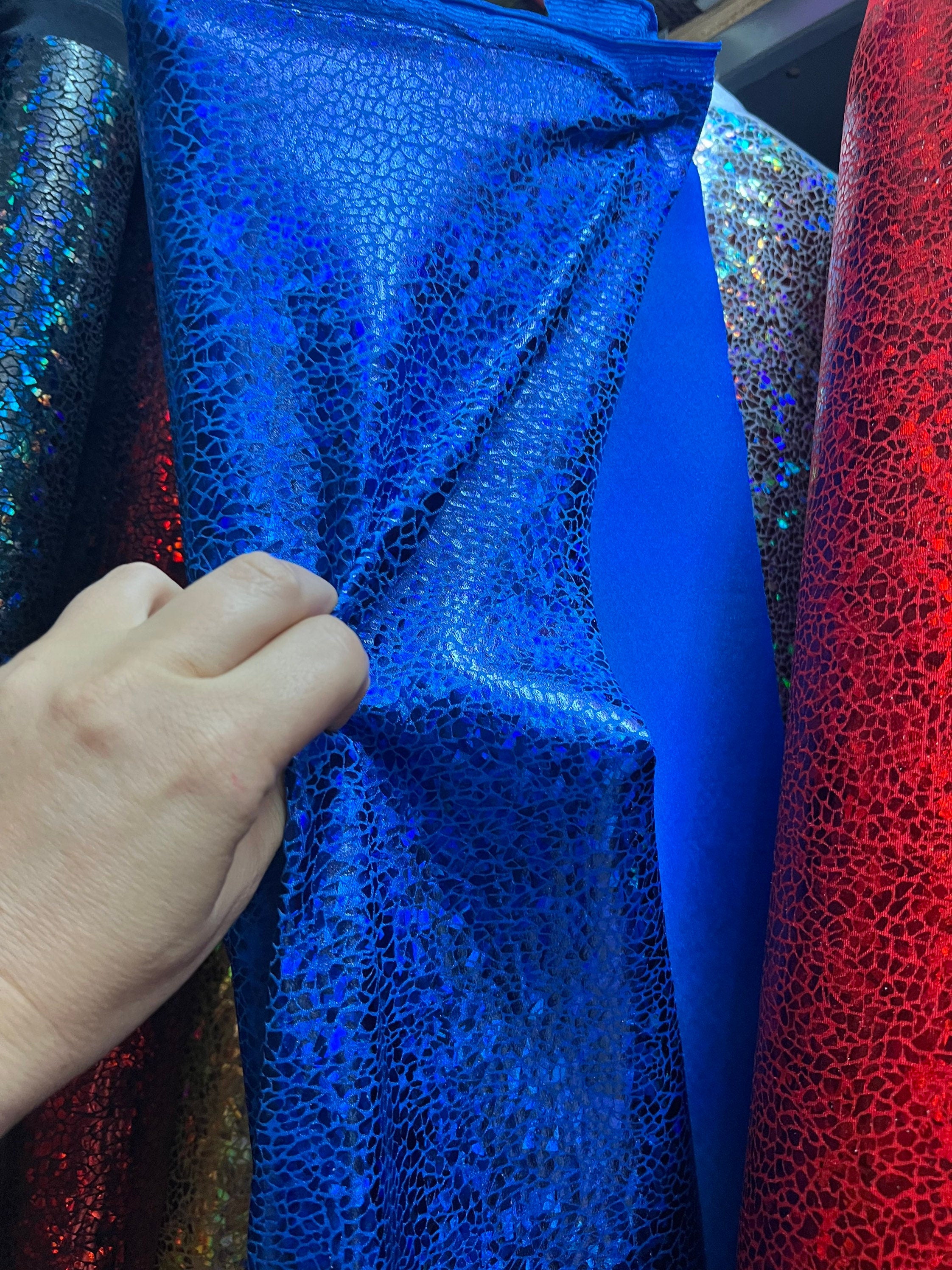 A hand holds a blue, cracked-patterned fabric resembling the Exotic Dancewear jumpsuit, reminiscent of an aerialist costume and surrounded by colorful fabrics.