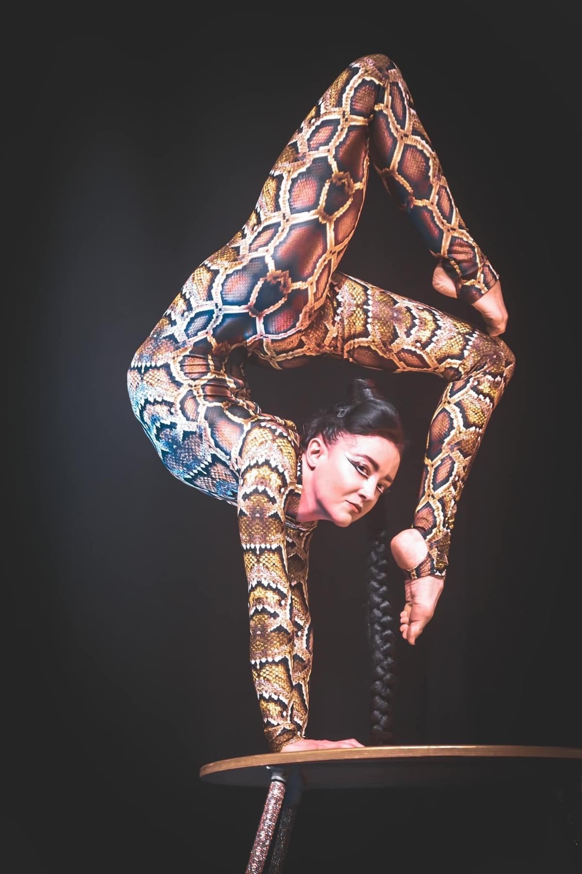 A contortionist balances on one hand, legs overhead, atop a round table against a dark background in an Elegant Swimwear Lycra Catsuit, where its 4-way stretch ensures every move is flawlessly executed.